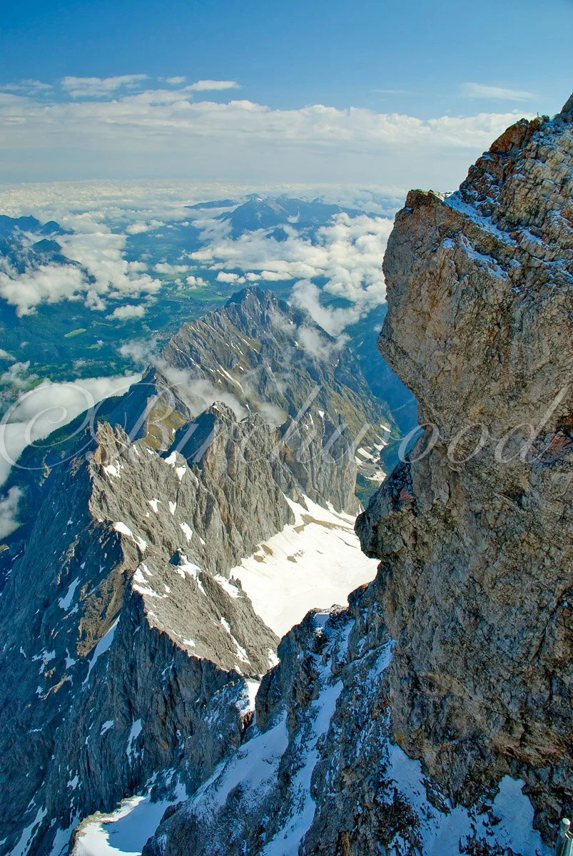 Aerial view of rugged snow-capped mountains with rocky cliffs, surrounded by clouds and a blue sky.