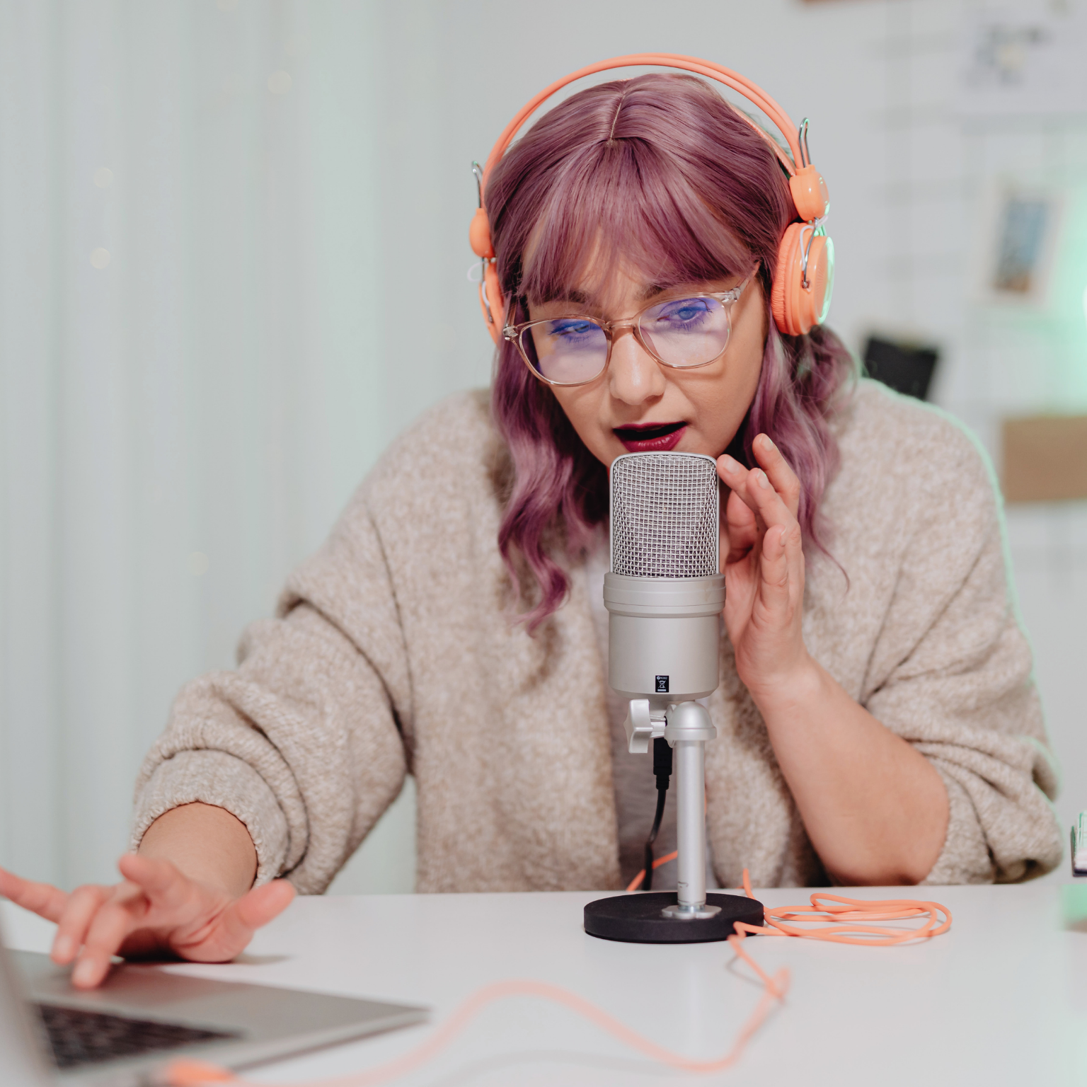 Young woman with pink hair, pink glasses, and orange headphones speaking into a podcast microphone