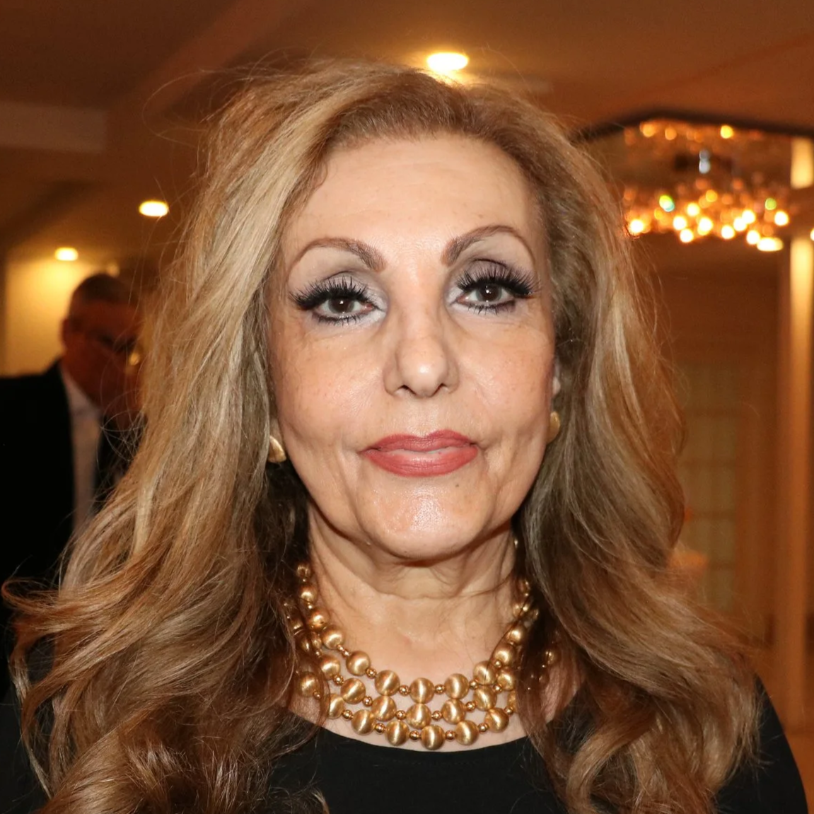 A woman with long, wavy hair, wearing earrings and a pearl necklace, at an indoor event.