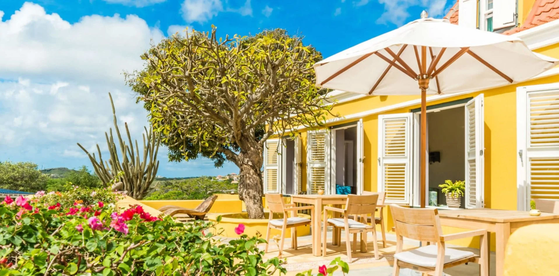 Outdoor patio with wooden furniture, umbrella, and yellow house with open shutters; surrounded by flowering plants, a tree, and a cactus under a blue sky.