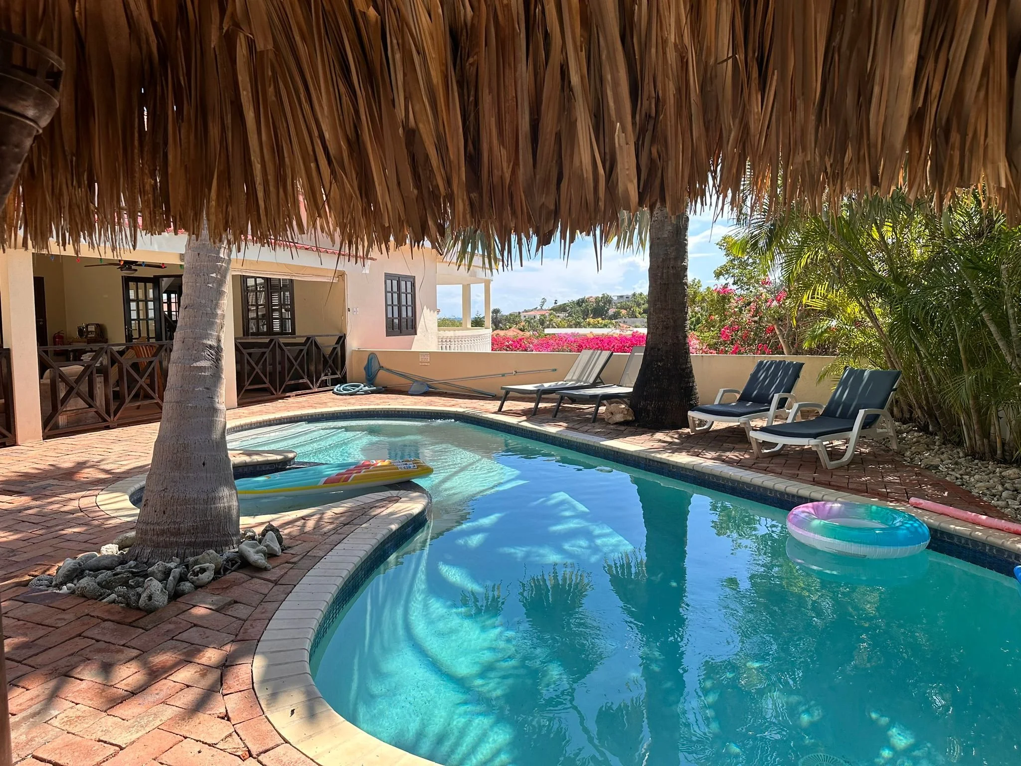 Tropical pool area with lounge chairs and palm trees, featuring a thatched roof canopy, vibrant flowers, and a tranquil swimming pool with floating rings.