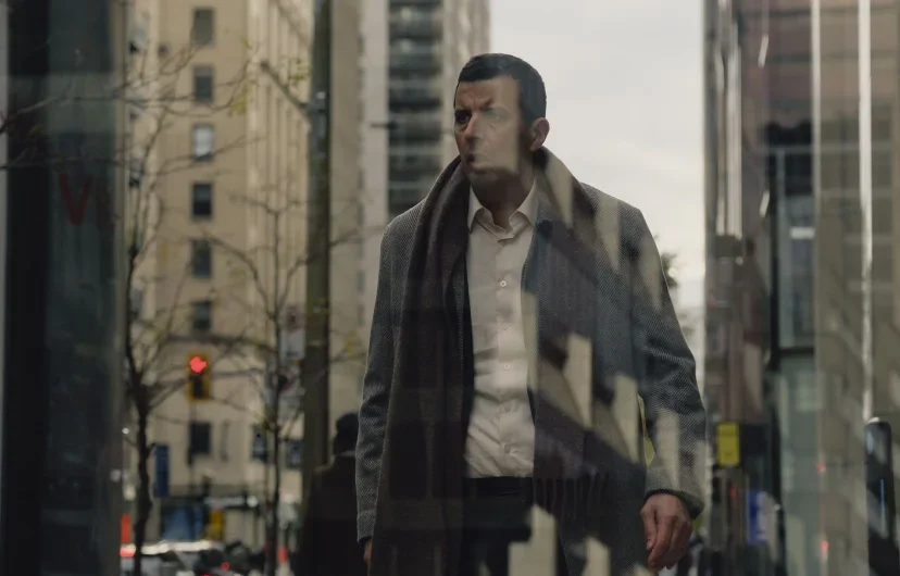 Homme dans une ville moderne, portant un manteau et une écharpe, vu à travers une vitre avec reflet.