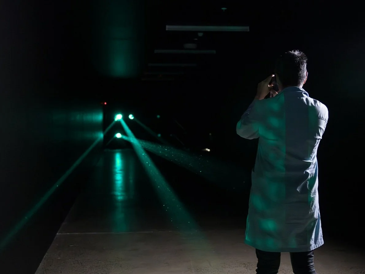 A man in a white coat stands in a dark room with a viewer, surrounded by laser beams and lights.
