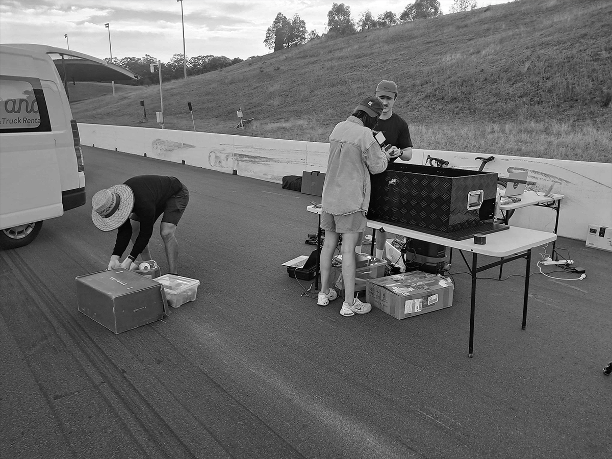 Three people working on a racetrack, two standing at a table with equipment and one bending over a box on the ground, with a van parked nearby.