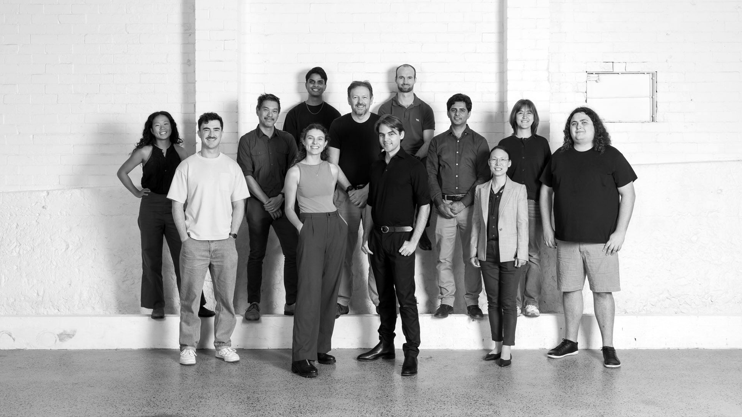 A diverse group of fifteen people standing together in a modern, minimalistic room with a white brick wall in the background.