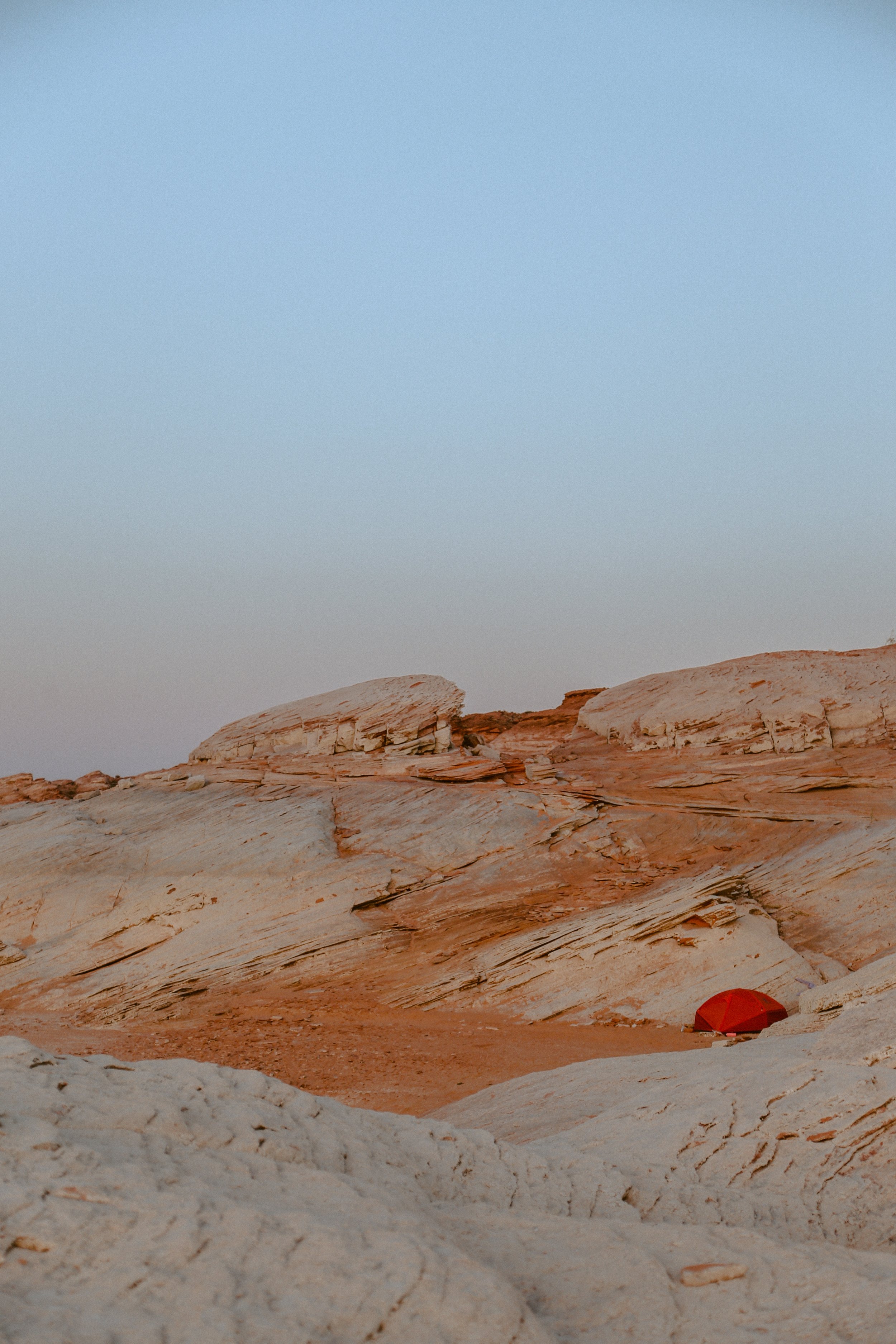A rugged desert landscape with layered sandstone formations and a small red tent in the distance, under a clear sky.