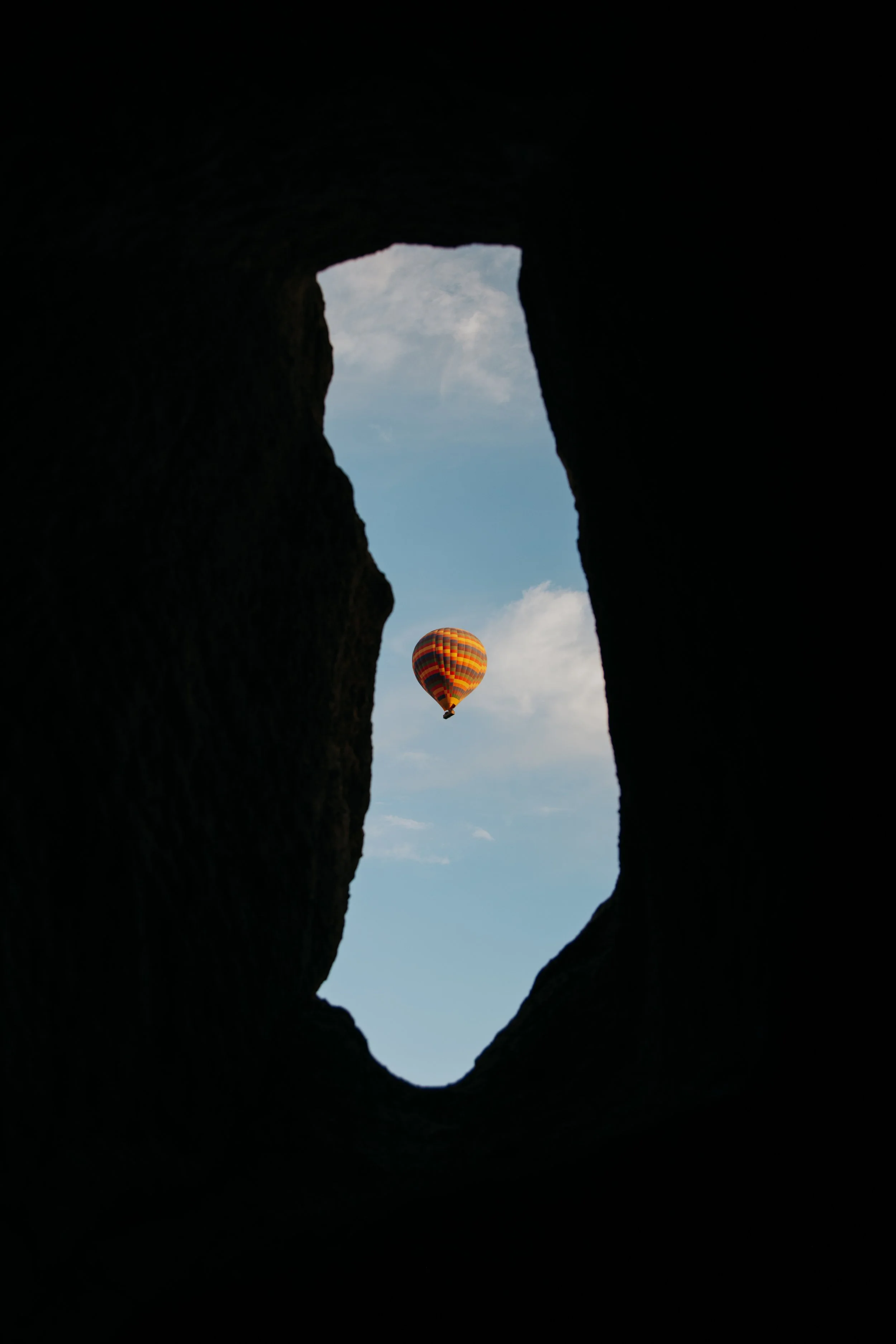 View through a dark cave or crevice showing a colorful hot air balloon floating in a blue sky with scattered clouds.