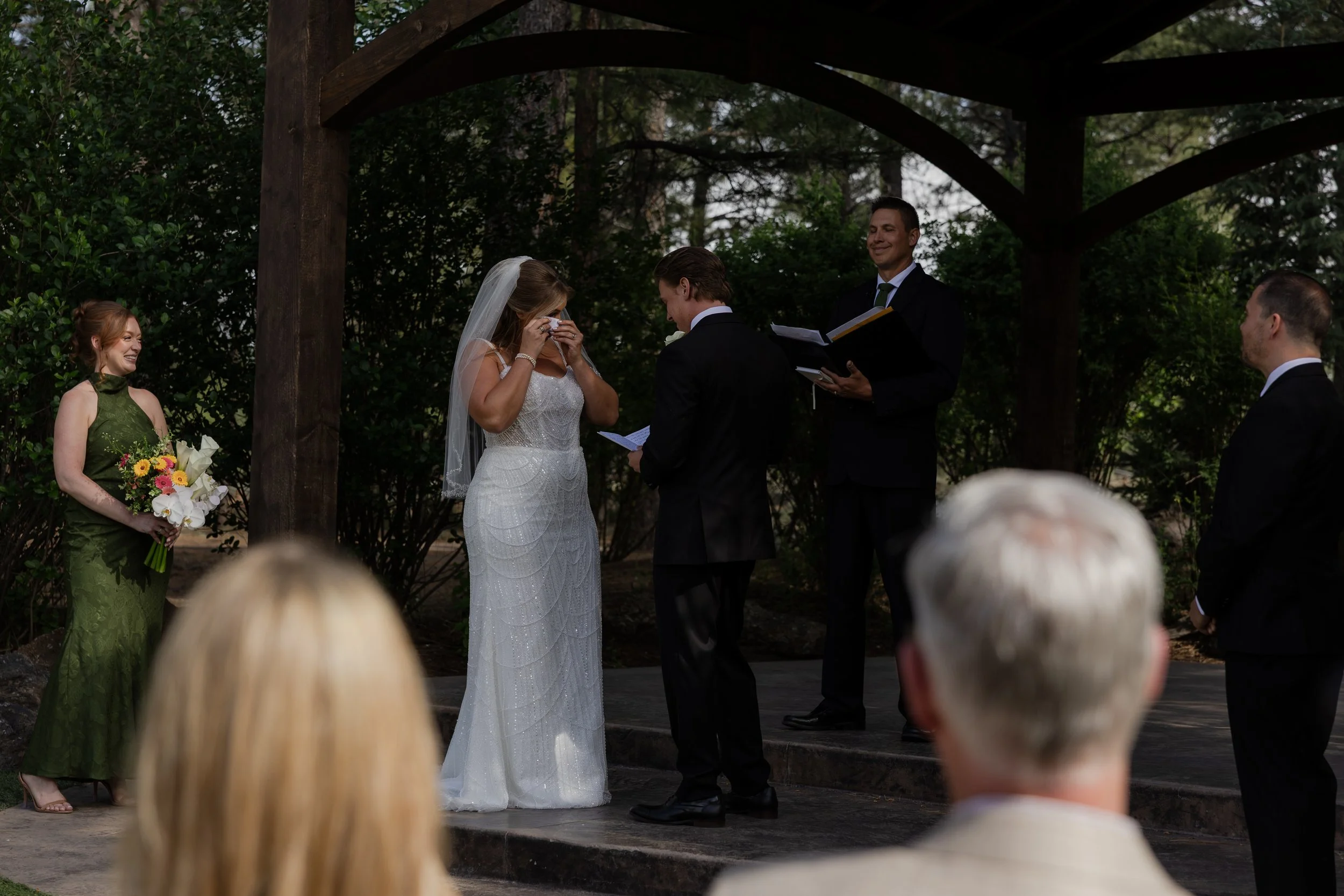 A bride and groom exchanging vows during an outdoor wedding ceremony, with bridesmaids and groomsmen standing nearby, and guests seated in front.