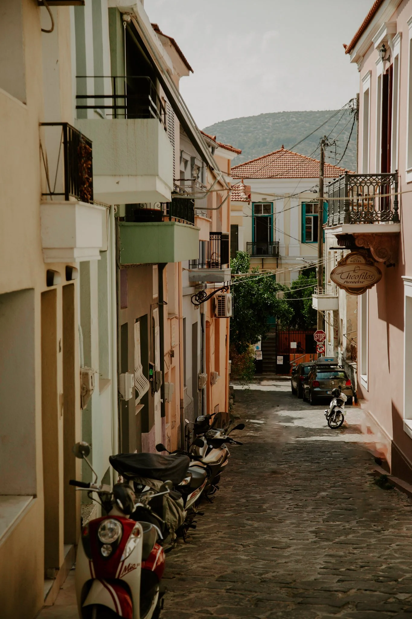A narrow cobblestone street lined with multicolored buildings, parked scooters, and cars; with a backdrop of hills and overcast sky.