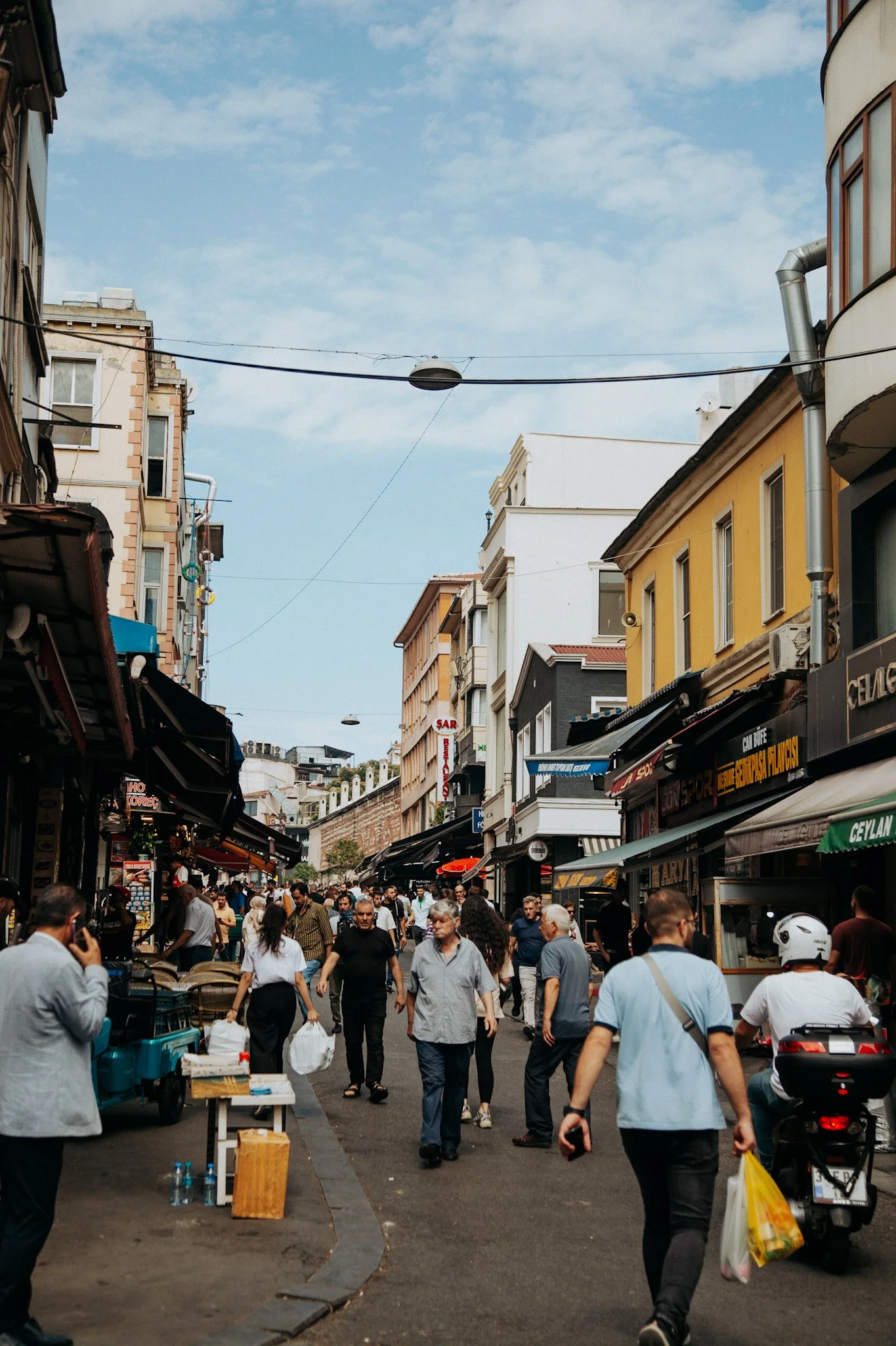 A busy city street with people walking, shops with awnings, and tall buildings under a partly cloudy sky.
