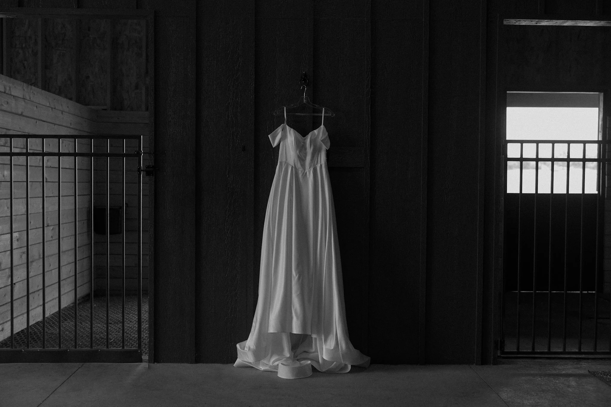 A white wedding dress hanging on a hanger inside a dark room with a wooden wall, iron railing, and a window in the background.