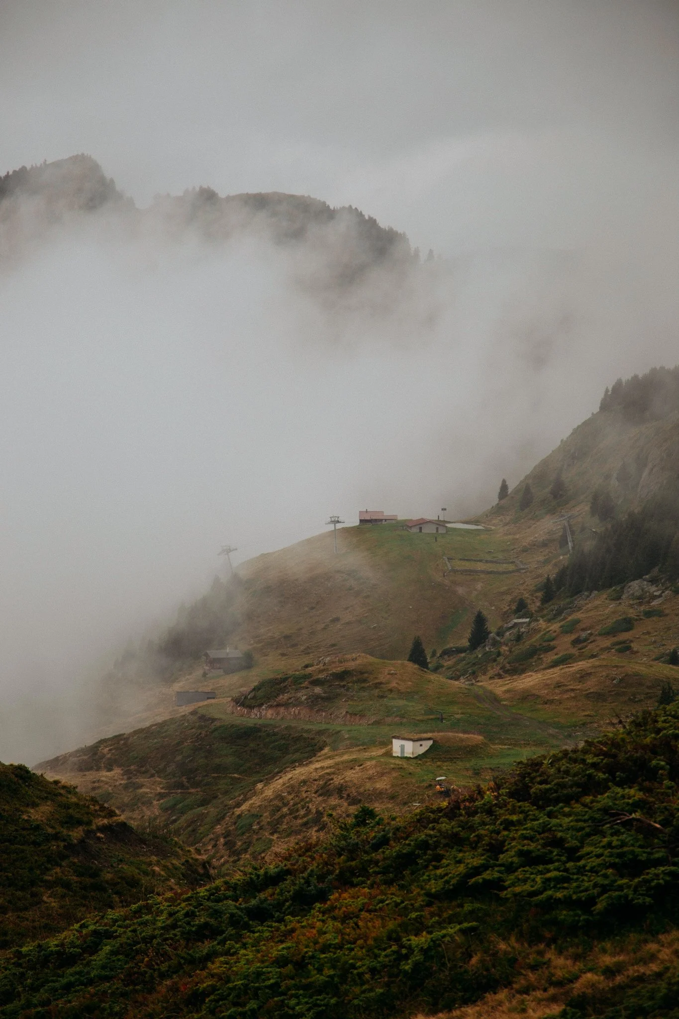 A foggy mountain landscape with green and brown slopes, scattered small buildings, and ski lift poles under a cloudy sky.