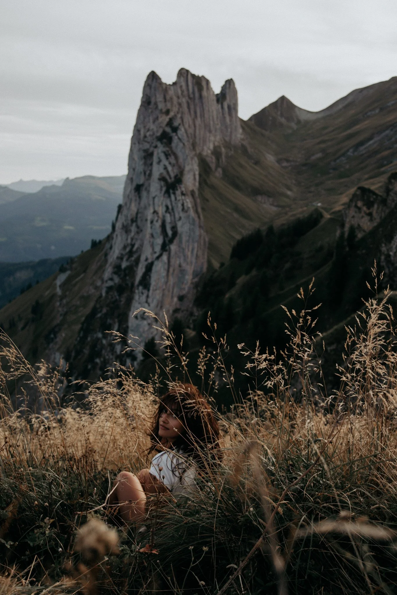 A woman sitting in tall grass in front of a mountainous landscape with a prominent rocky peak.