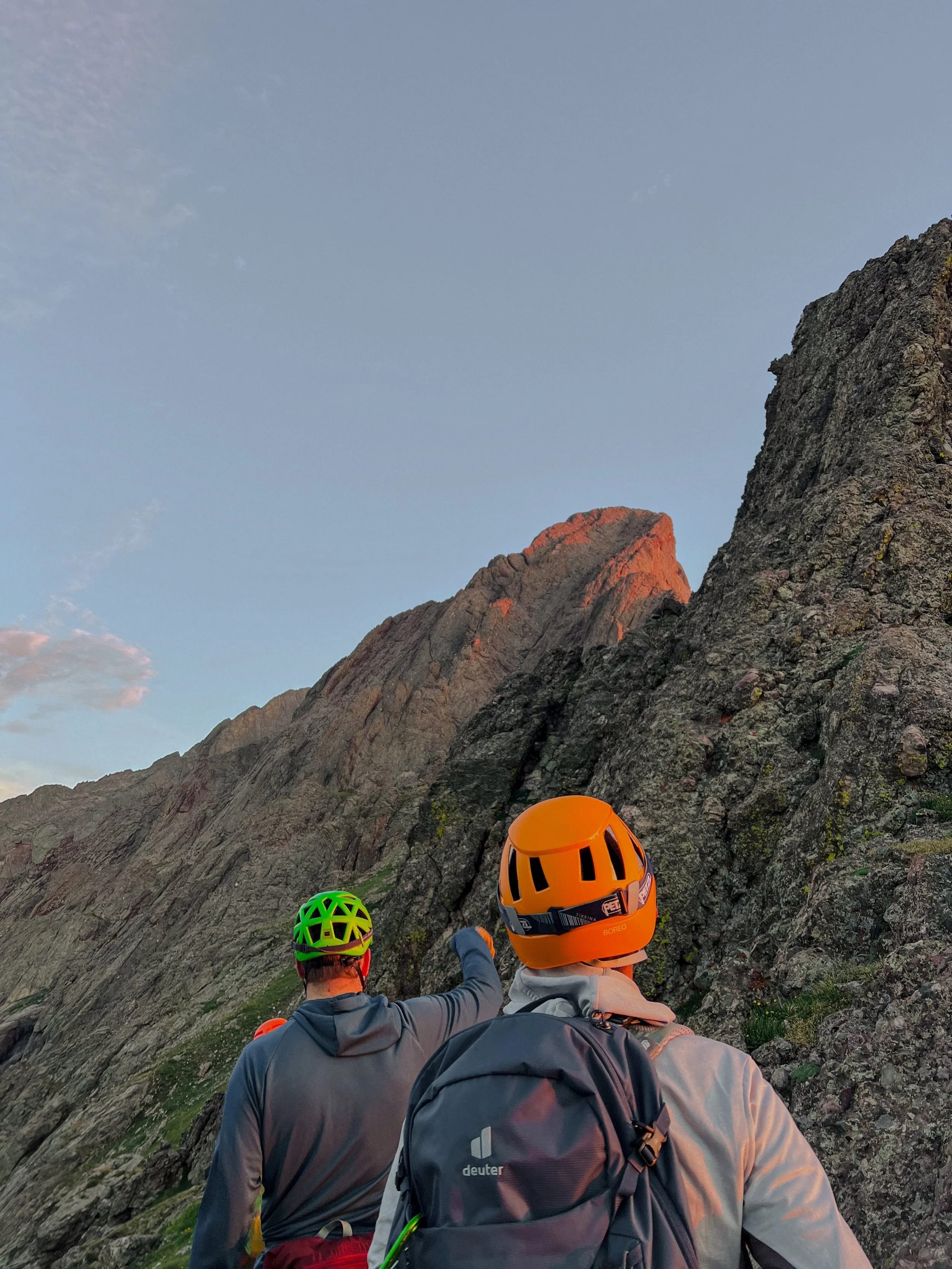 Two climbers with helmets and backpacks watching a large mountain cliff at sunset.