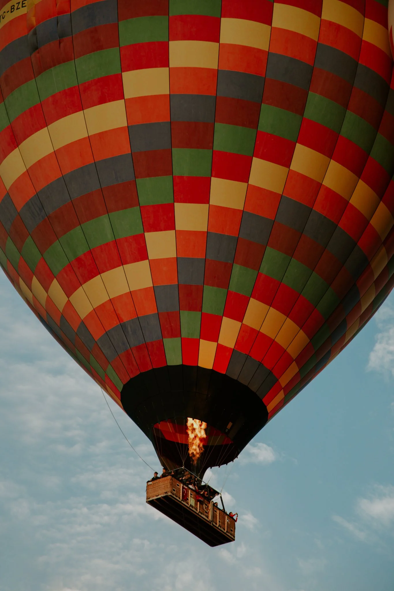 A hot air balloon with a colorful checkered pattern of red, yellow, green, and black panels, with a flame visible inside the balloon and a wicker basket carrying passengers, floating in a partly cloudy sky.