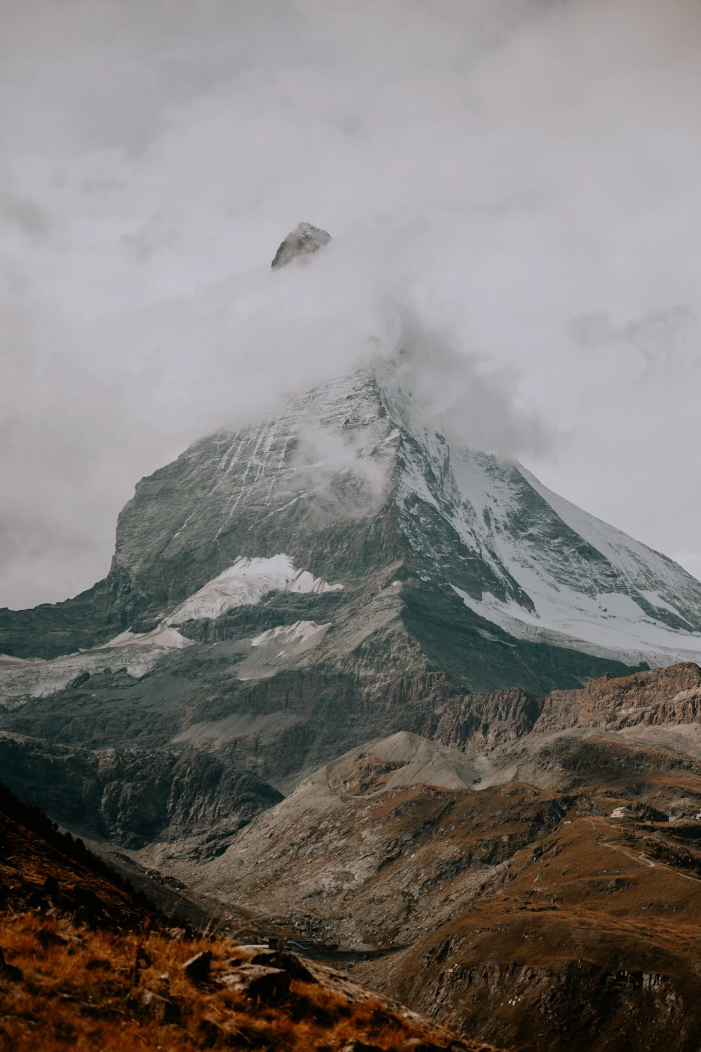 Snow-capped mountain peak partly obscured by clouds, with rugged terrain and grassy slopes in the foreground.