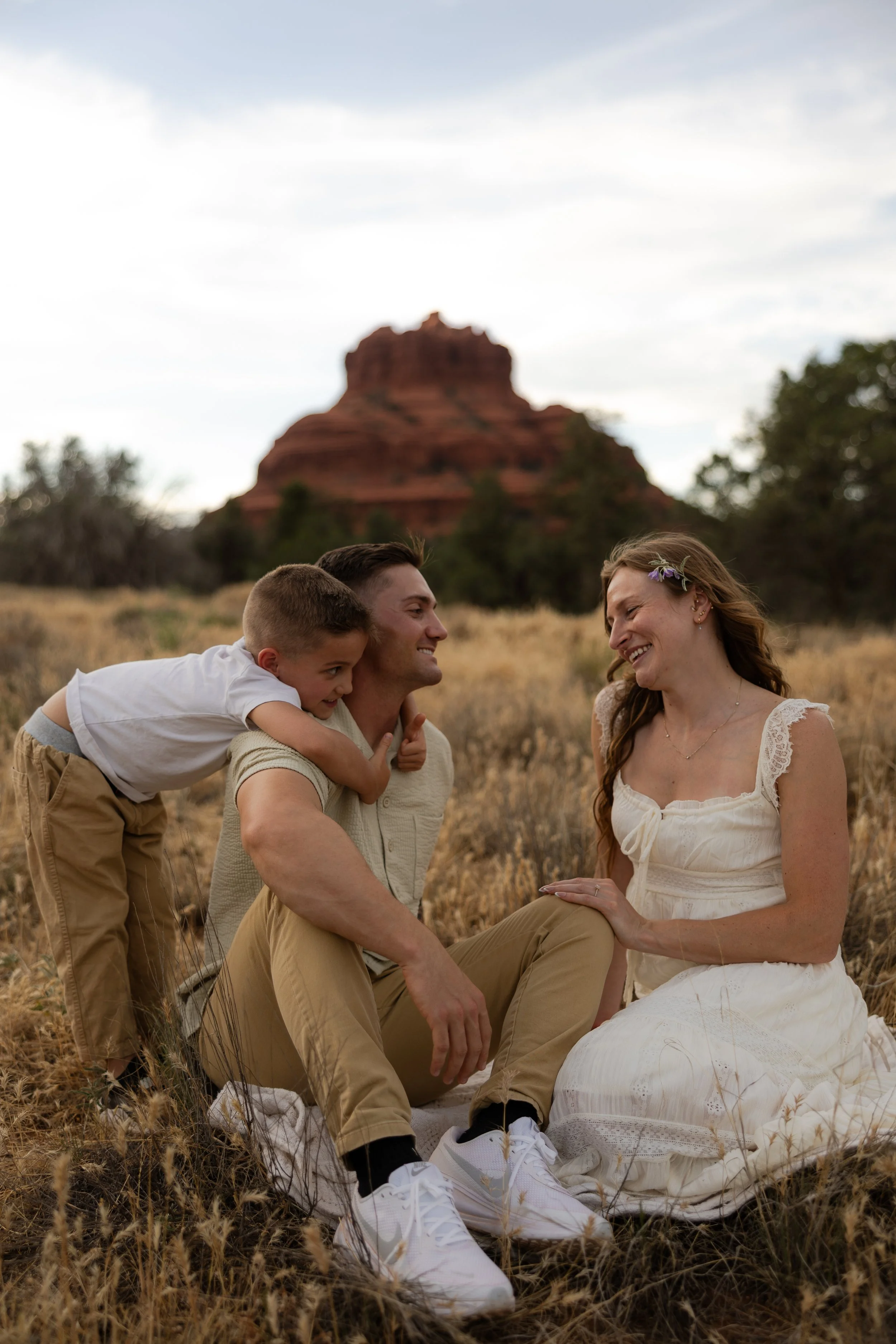 A family of three sitting in a field with dry grass, smiling and embracing each other, with a red rock formation in the background and a partly cloudy sky.