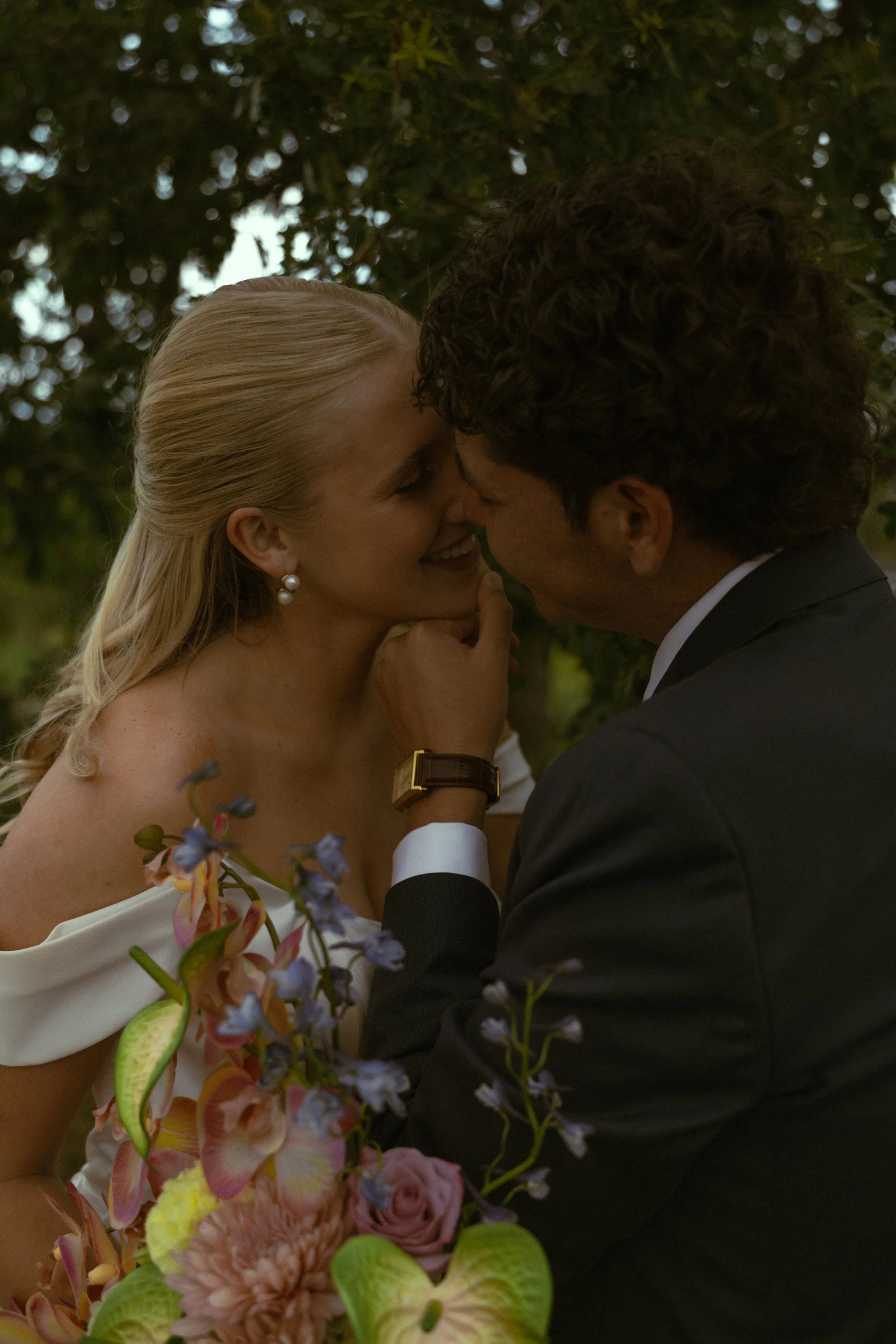 A couple sharing a kiss outdoors surrounded by trees, with the woman wearing pearl earrings and holding a colorful bouquet of flowers.
