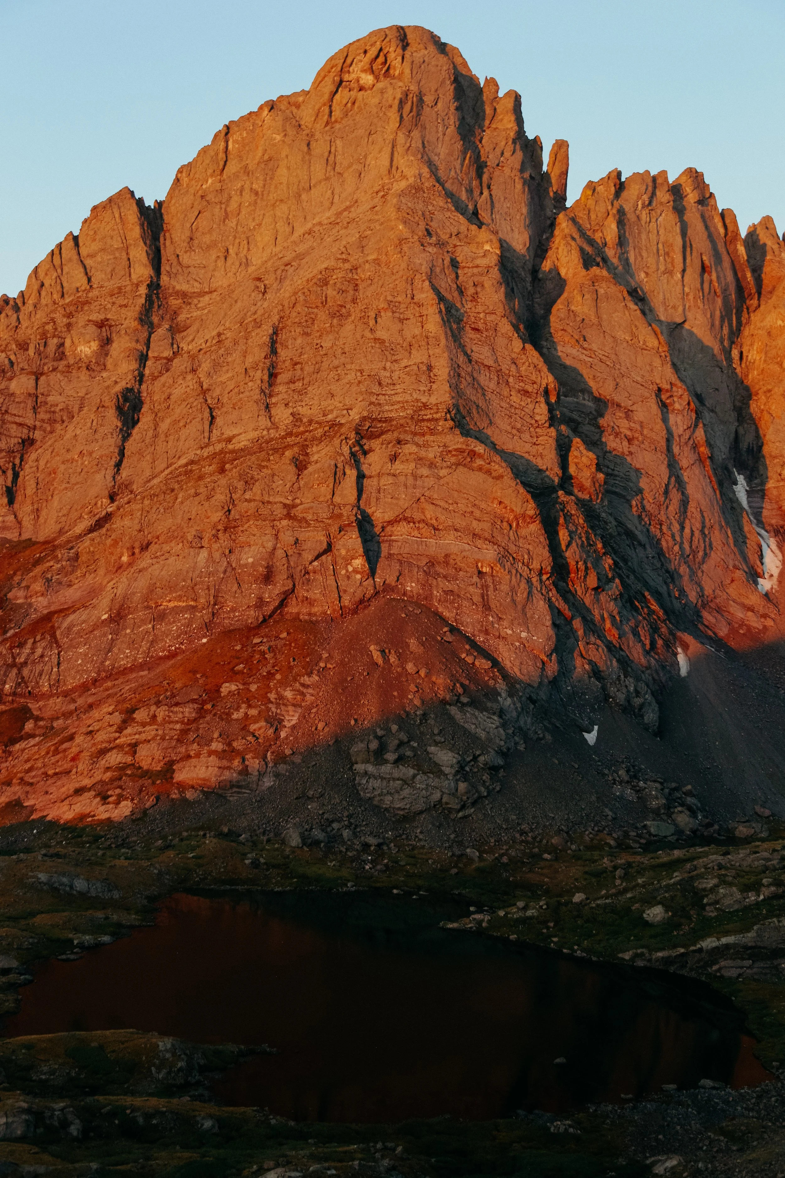 A large sandstone mountain illuminated by sunset light, with a small pond at its base and rocky terrain surrounding it.