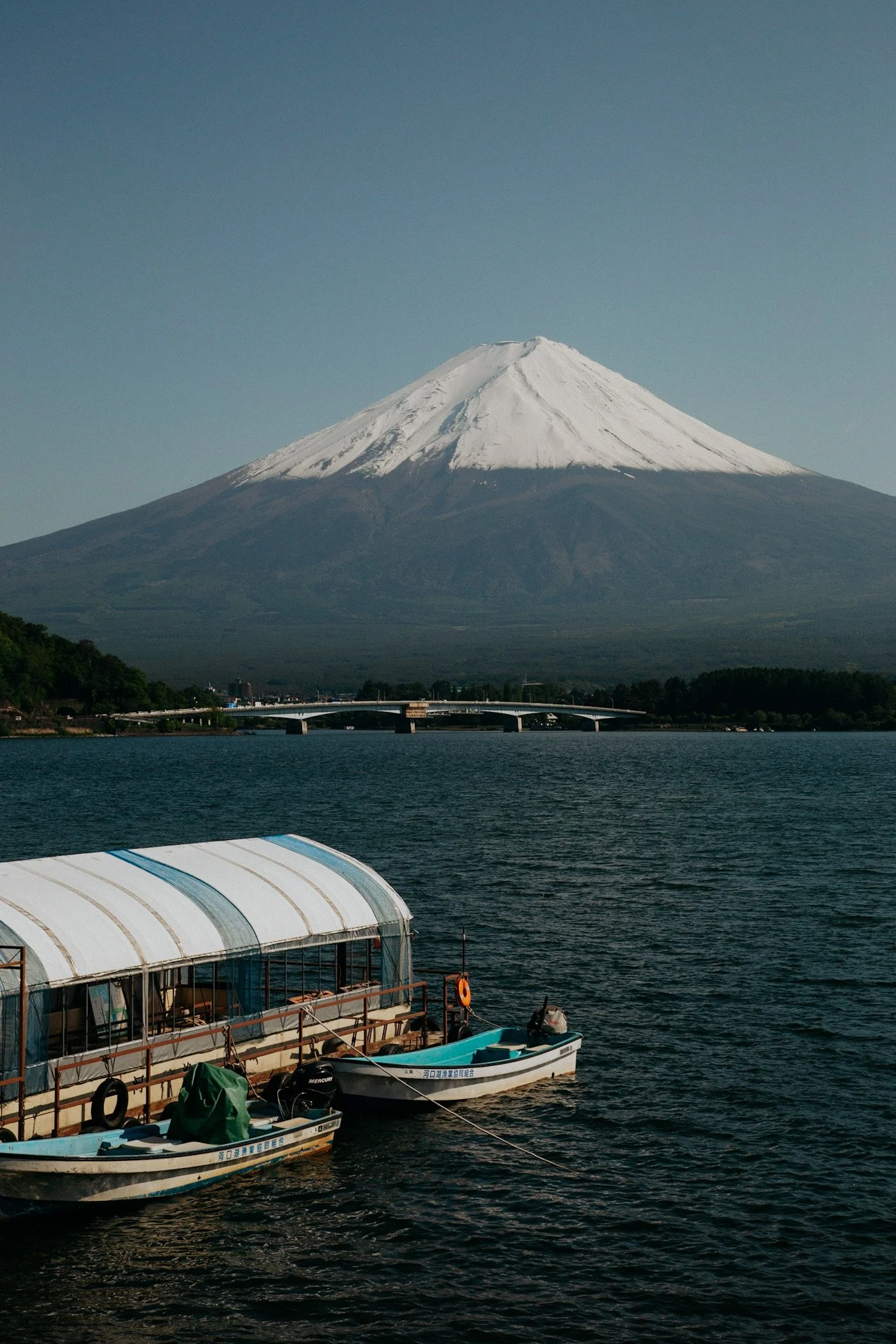 Snow-capped Mount Fuji in the background with boats floating on a lake in the foreground, and a bridge across the water.