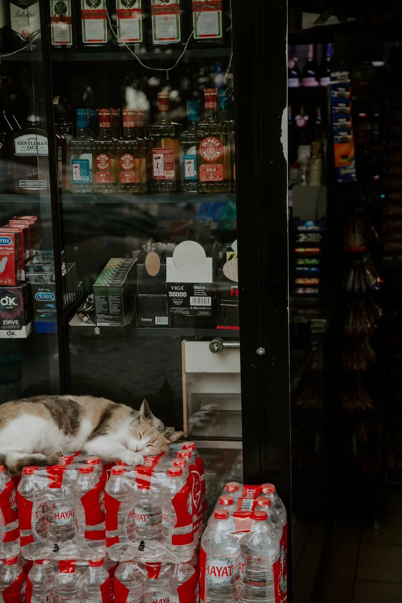 A cat sleeping on top of bottled water in a store display case behind glass, with shelves of various products inside and outside the case.