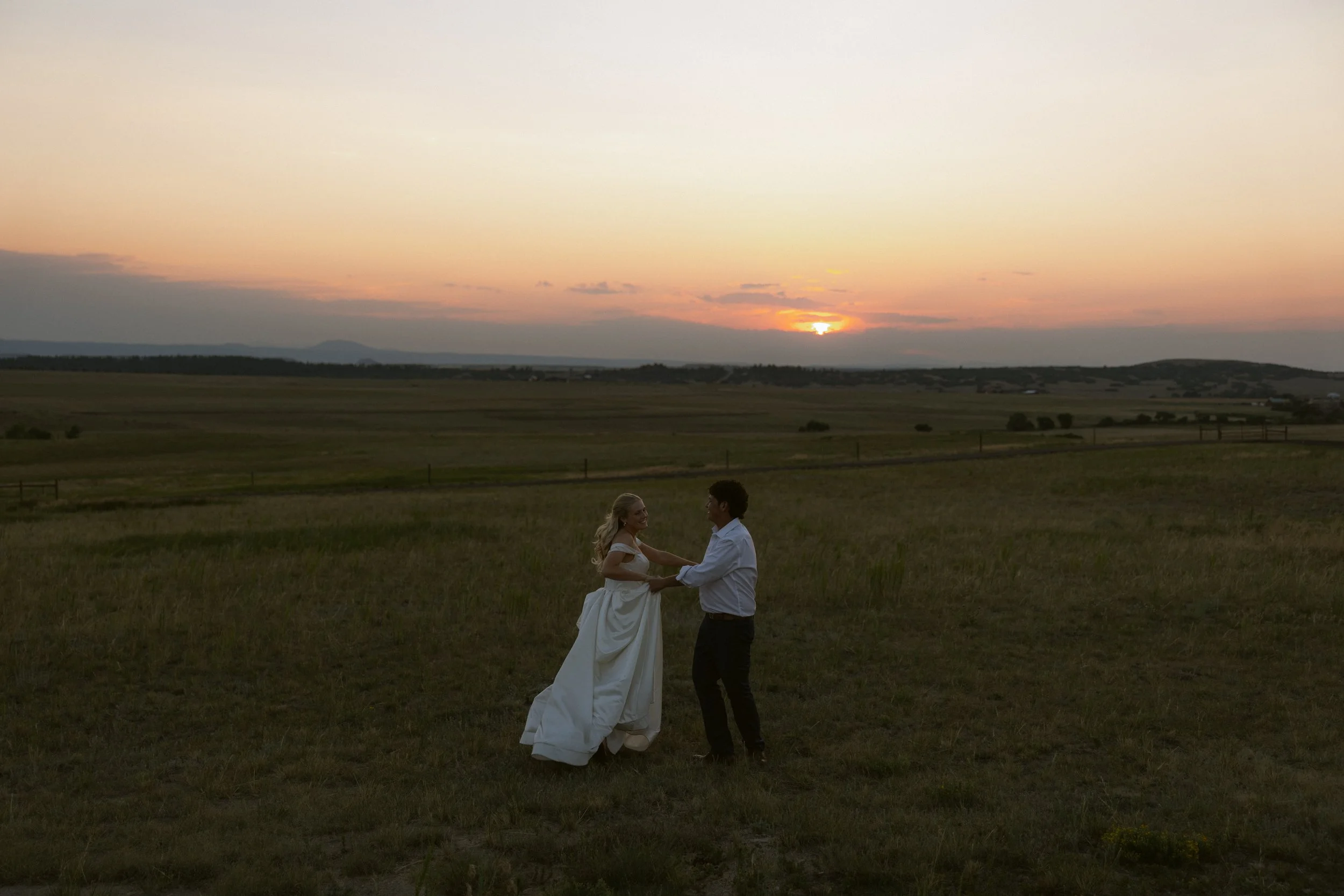 A couple holding hands and dancing in a field at sunset, with a scenic landscape and a cloudy sky.