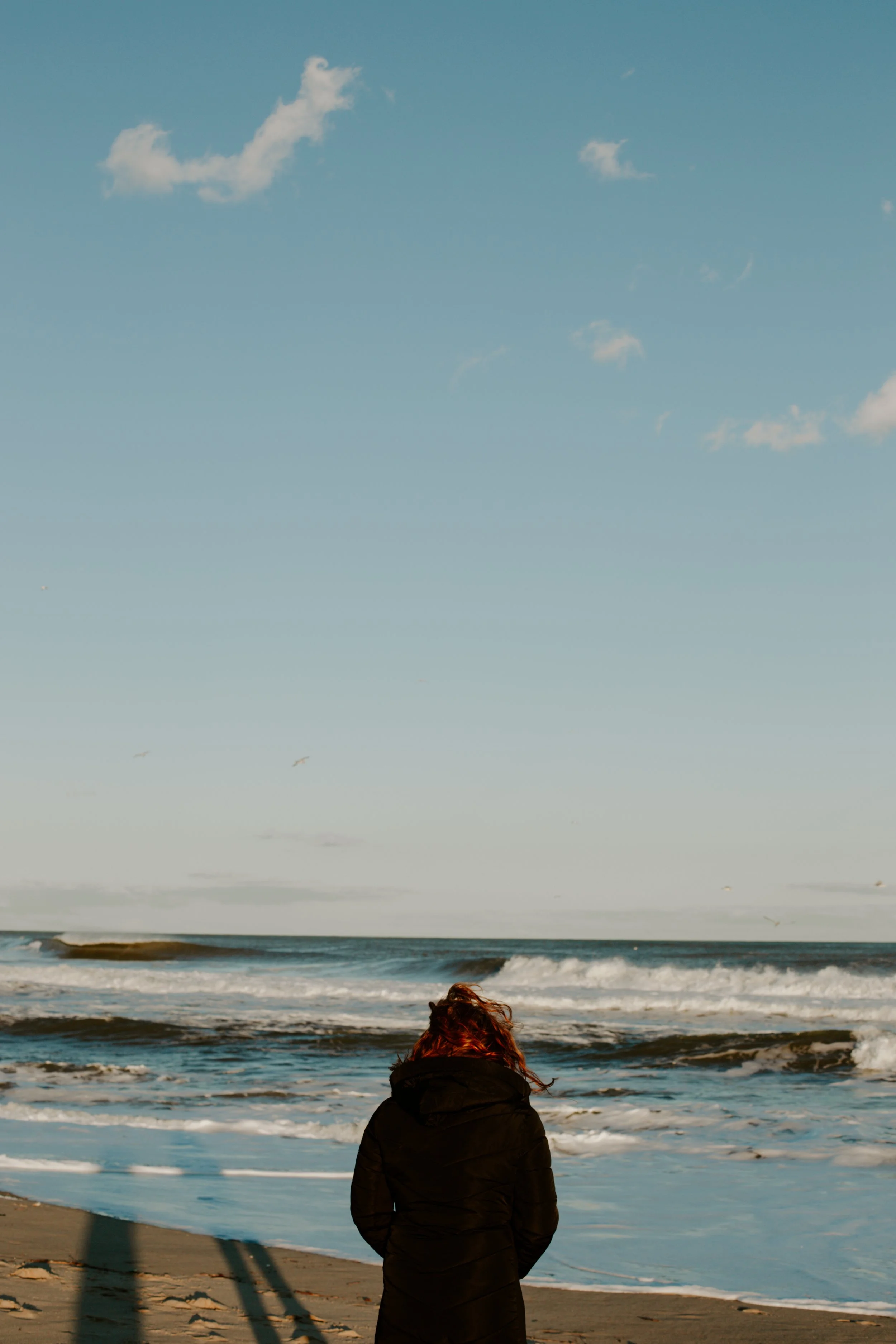 A woman with red hair wearing a black jacket standing on a sandy beach, looking at the ocean with waves, under a blue sky with a few clouds.