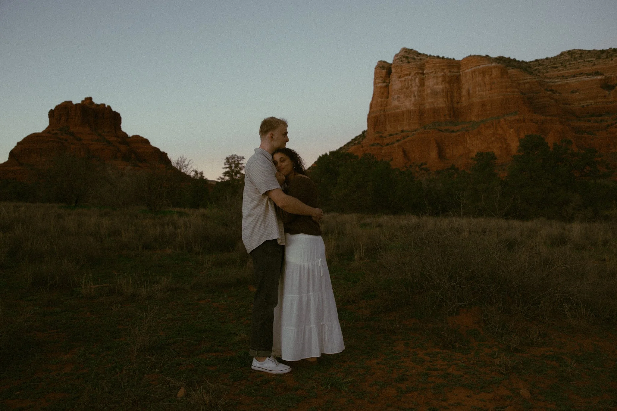 A couple embracing in a field with red rock formations and a clear sky in the background during sunset.