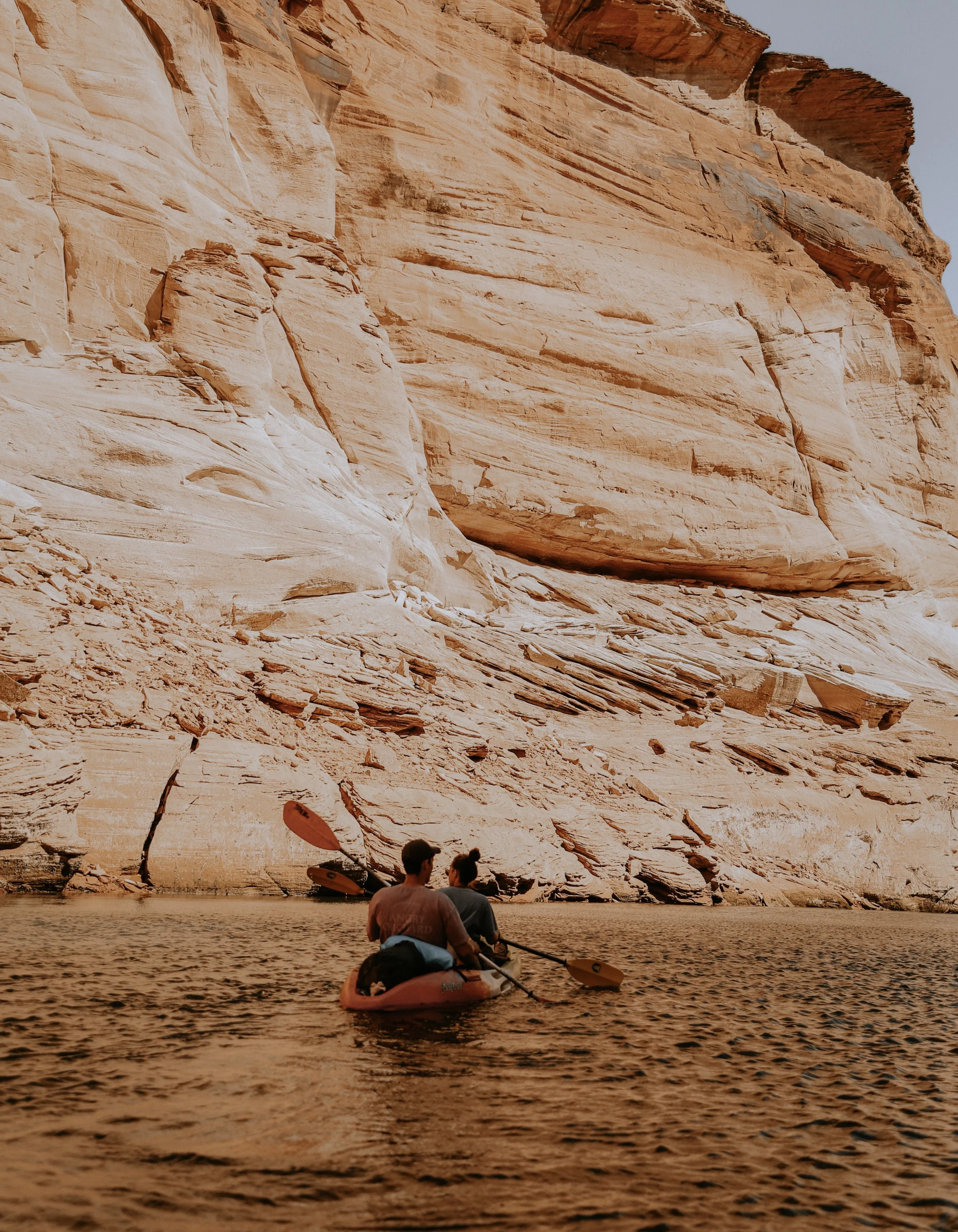 Two people in a kayak paddling in a body of water in front of a steep sandy-colored rock formation.