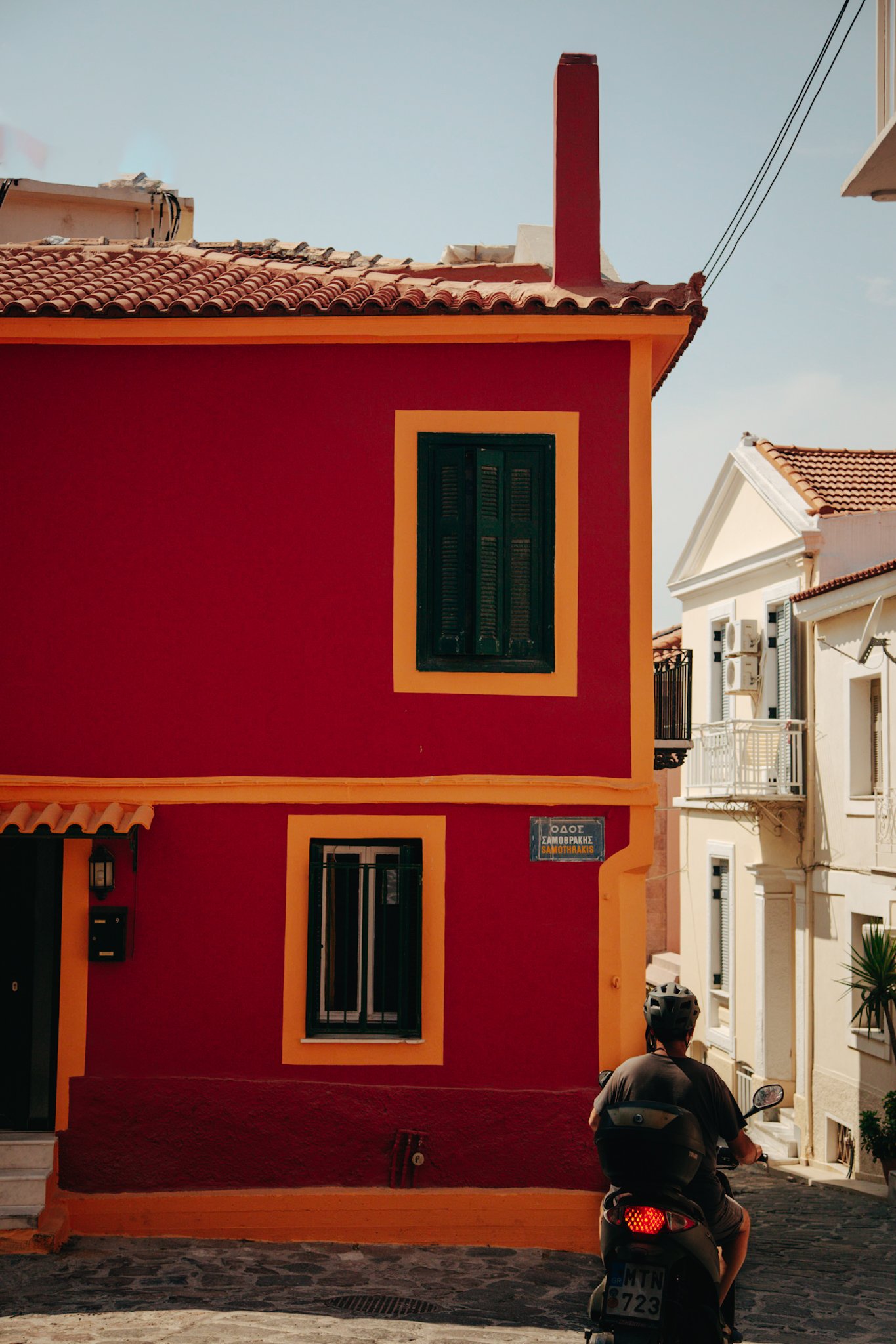 A man riding a scooter past a colorful red and yellow building on a cobblestone street, with white buildings nearby under a clear sky.
