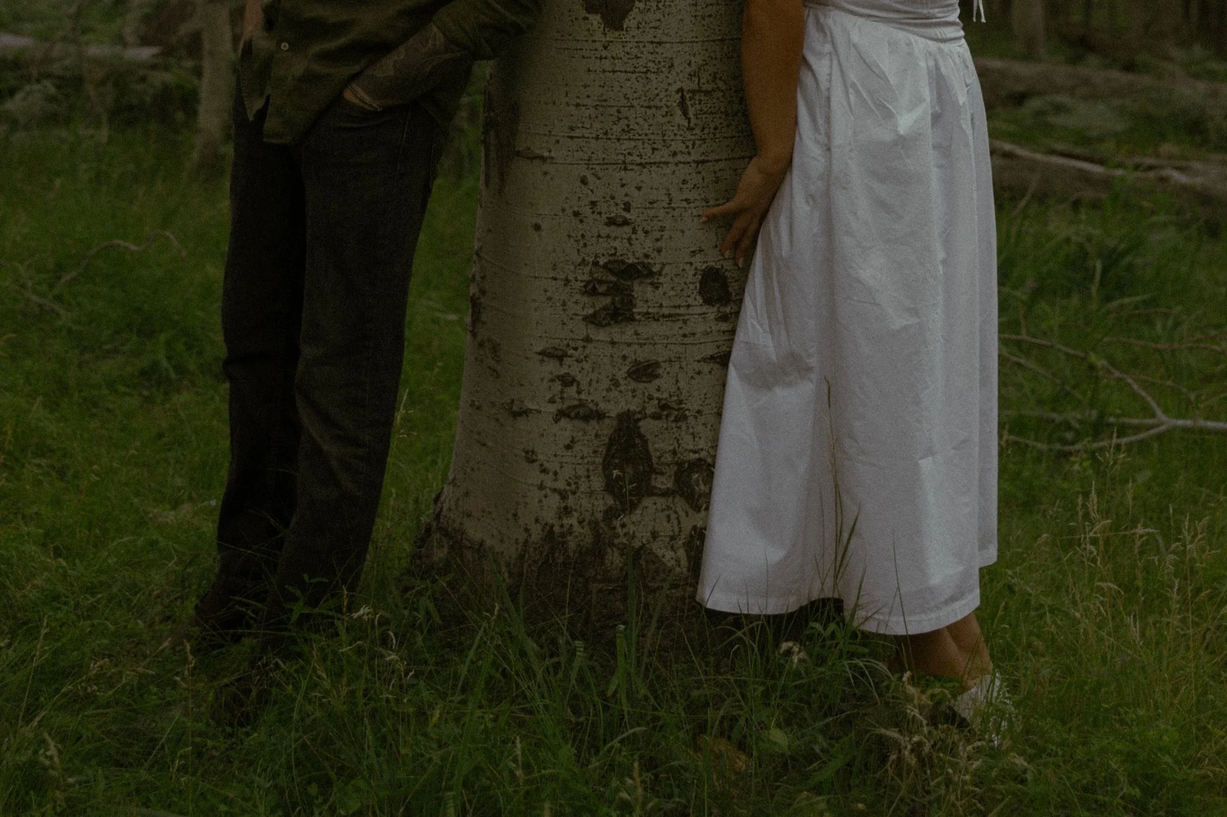 Two people, one dressed in white with a white skirt and the other in dark clothing, are standing on either side of a large tree trunk in a forested area. Their hands are resting on the tree.
