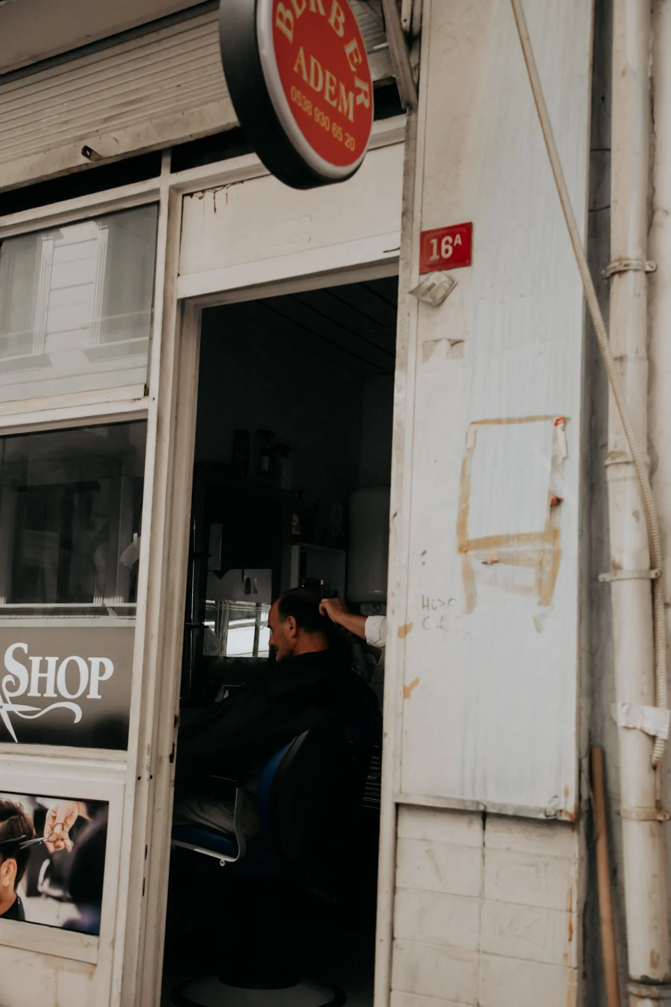 A man sitting inside a barbershop, getting a haircut, viewed through the shop's window and door, with signs and a red street number 16A outside.