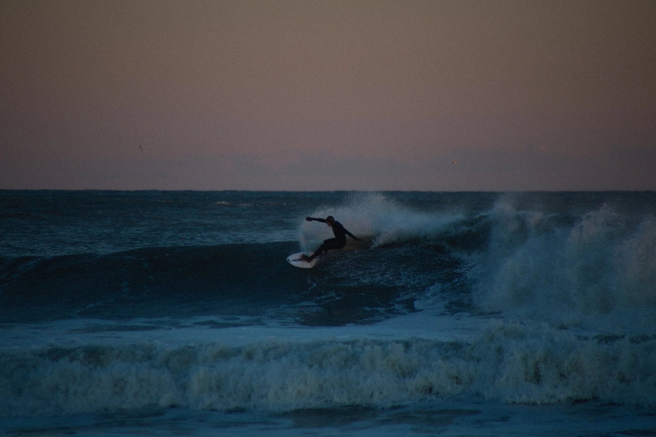 A person surfing on a wave in the ocean during dusk or dawn with a pinkish sky in the background.
