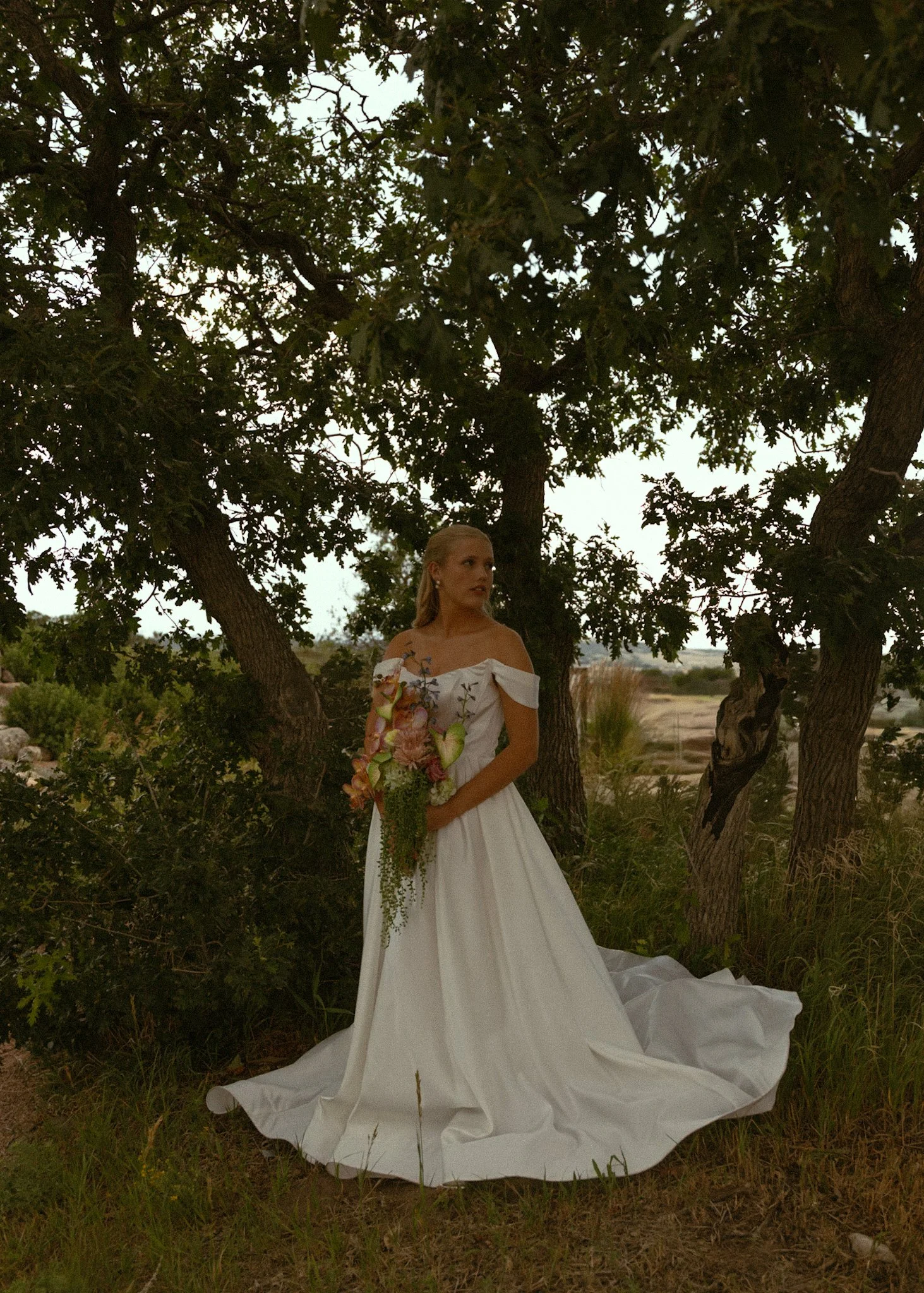 A woman in a white off-shoulder dress holding a bouquet of flowers standing under trees outdoors during dusk.