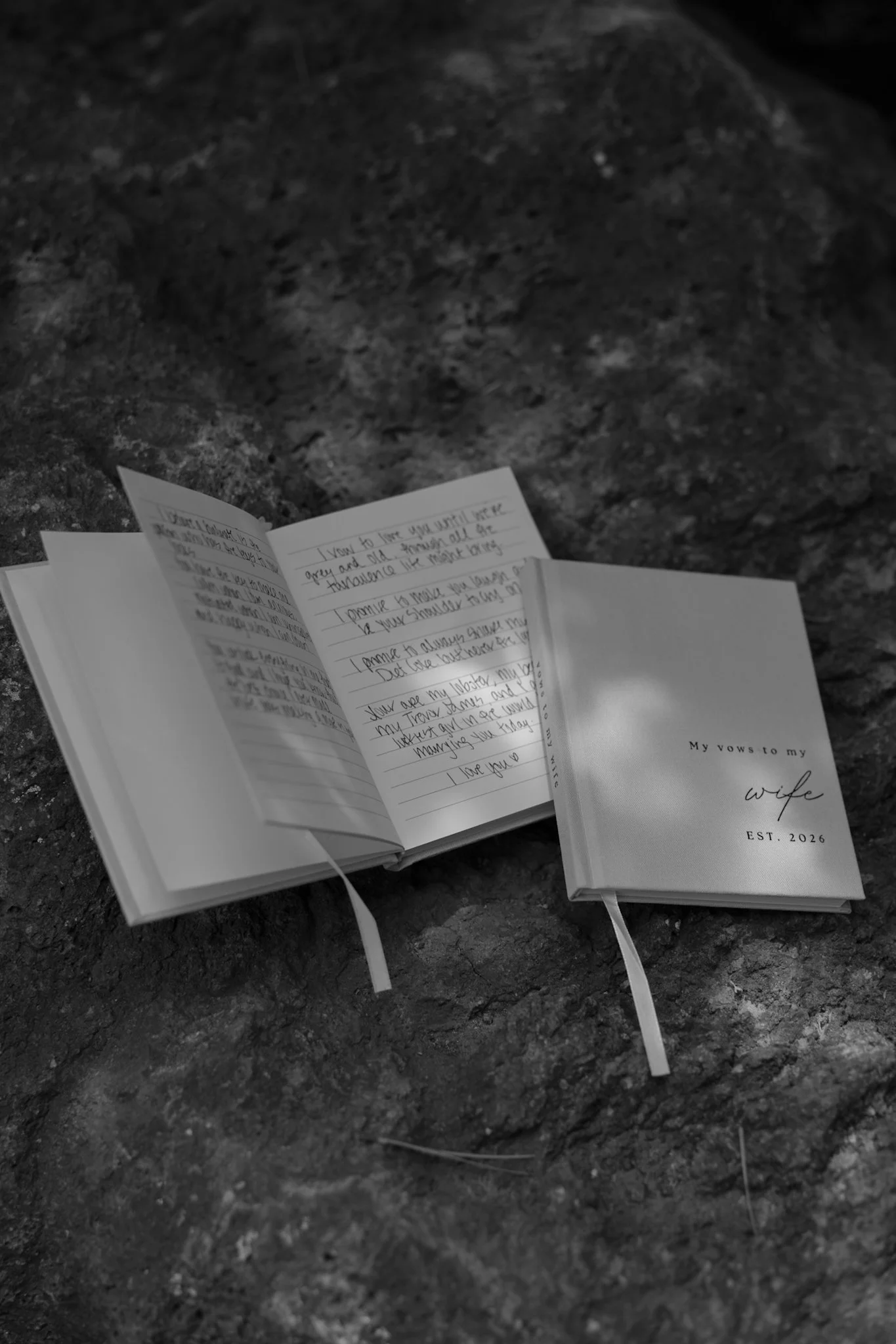 An open notebook with handwritten text and a closed book titled 'My vows to my wife, EST. 2026' resting on a rocky surface.