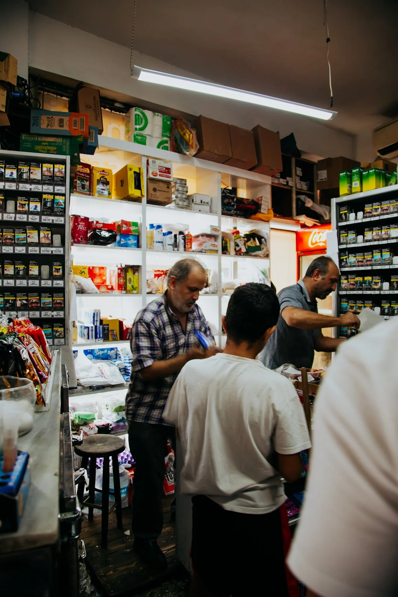 Inside a small store with shelves stocked with various products, three men are working behind the counter and a customer is in front of the counter.