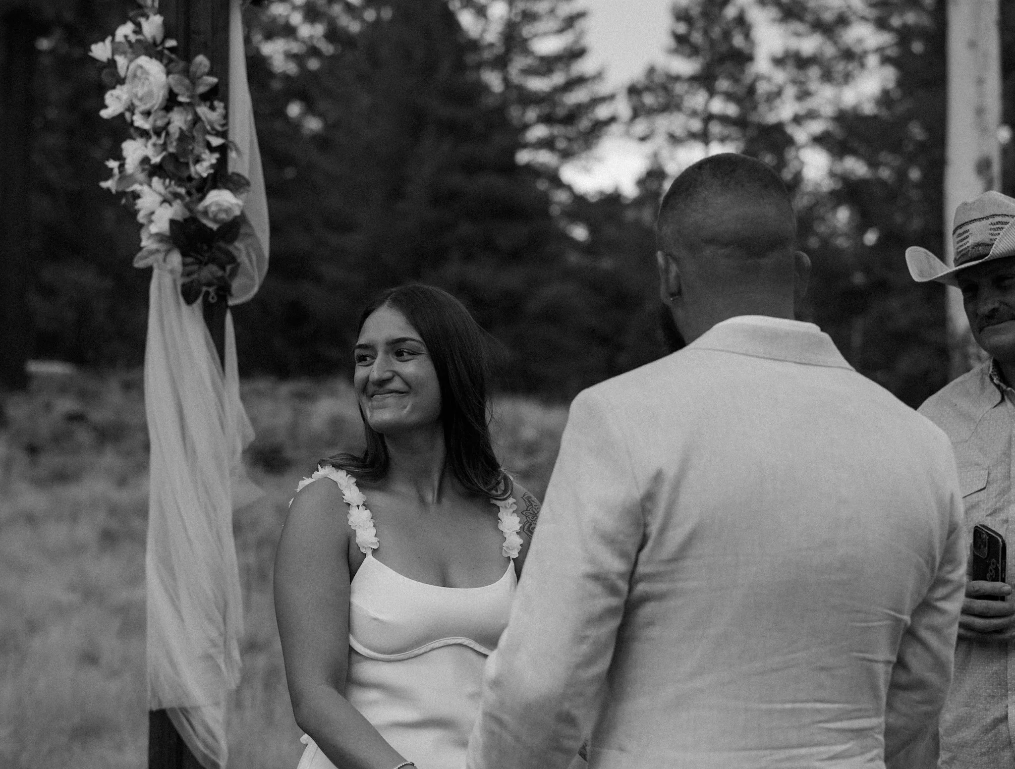 A woman in a white dress with floral straps smiling at a man in a white suit during an outdoor wedding ceremony, with another man holding a phone and a decorated flag in the background, and trees surrounding the scene.