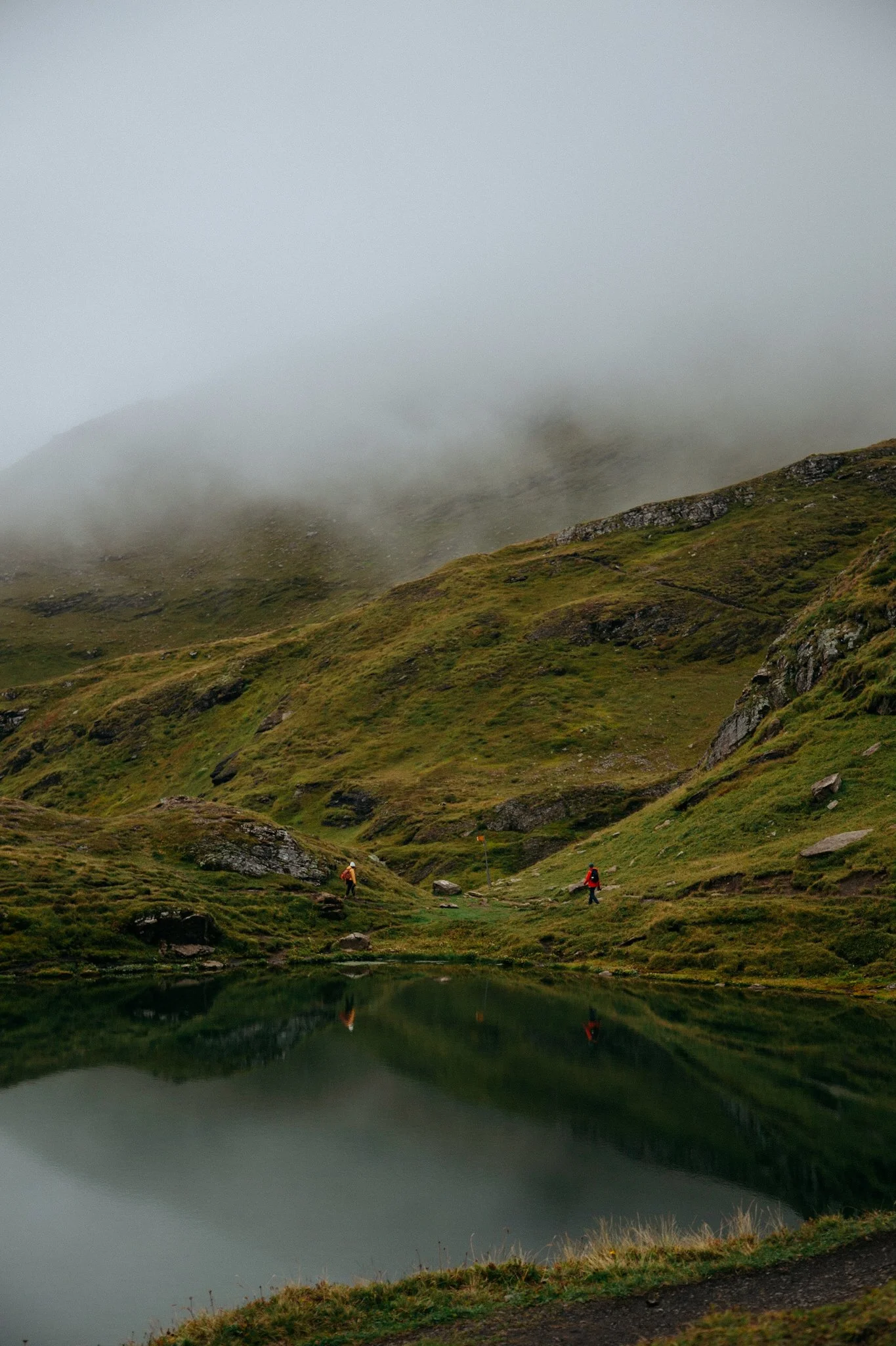 Two hikers walking along a trail near a small lake in a foggy, green mountainous landscape.
