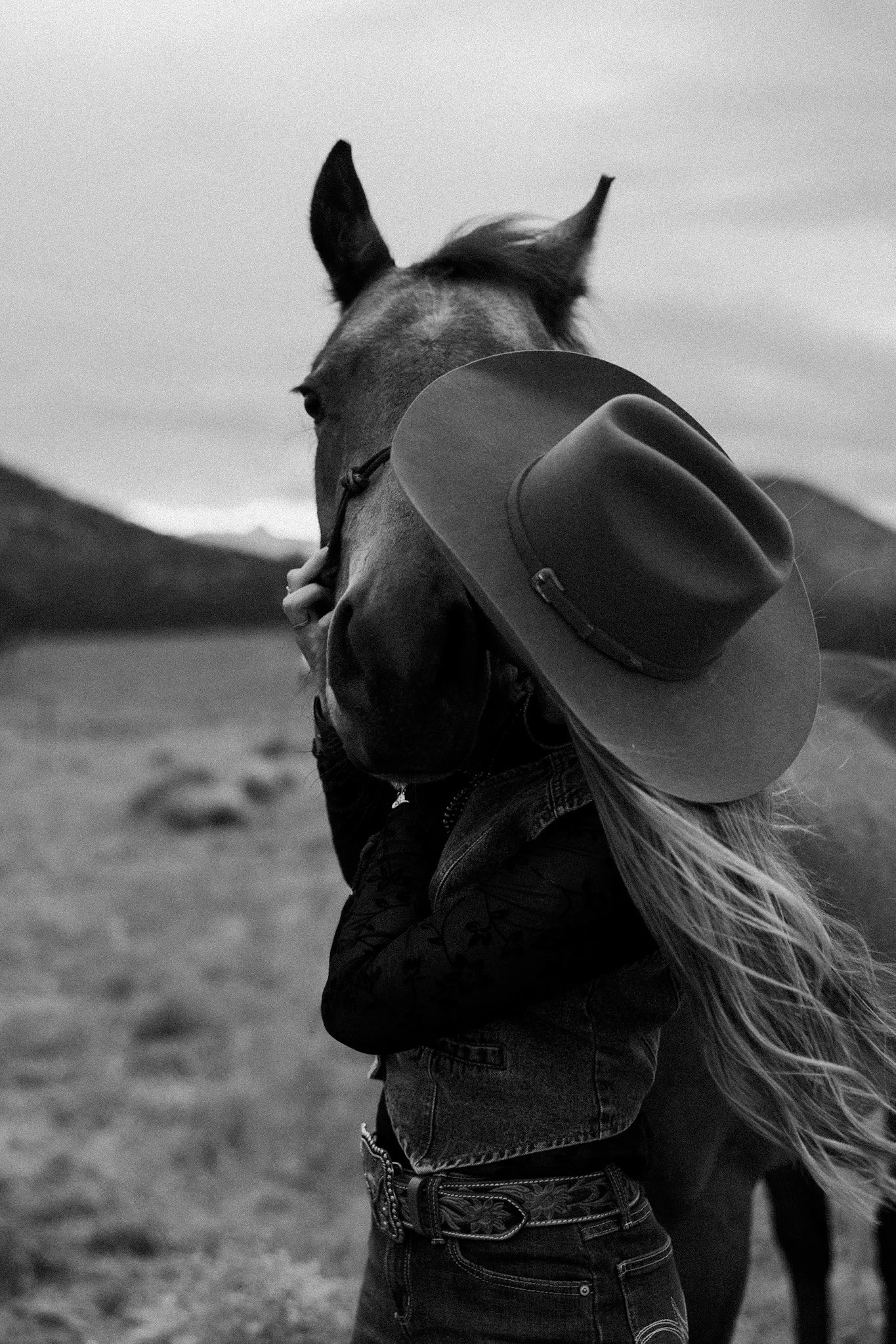 A woman in a cowboy hat and denim jacket hugging a horse outdoors, mountains in the background, black and white photo.