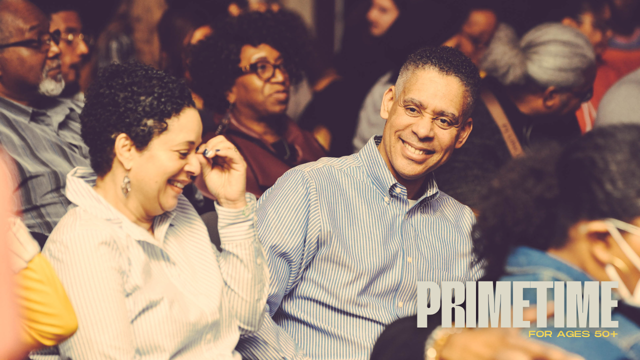 Audience members at a small group event called Primetime, smiling and engaging; one man in the foreground is looking at the camera with a big smile, wearing a striped shirt.