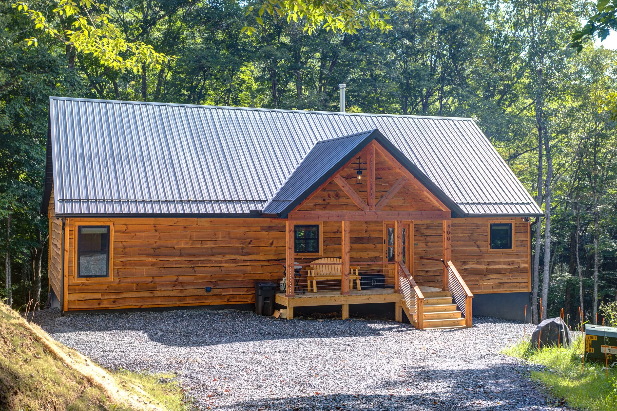 A beautiful rustic mountain cabin surrounded by peaceful woodland, featuring warm natural-wood siding, a durable metal roof, black-framed windows, and a welcoming covered front porch with exposed timber details. The perfect combination of country cha