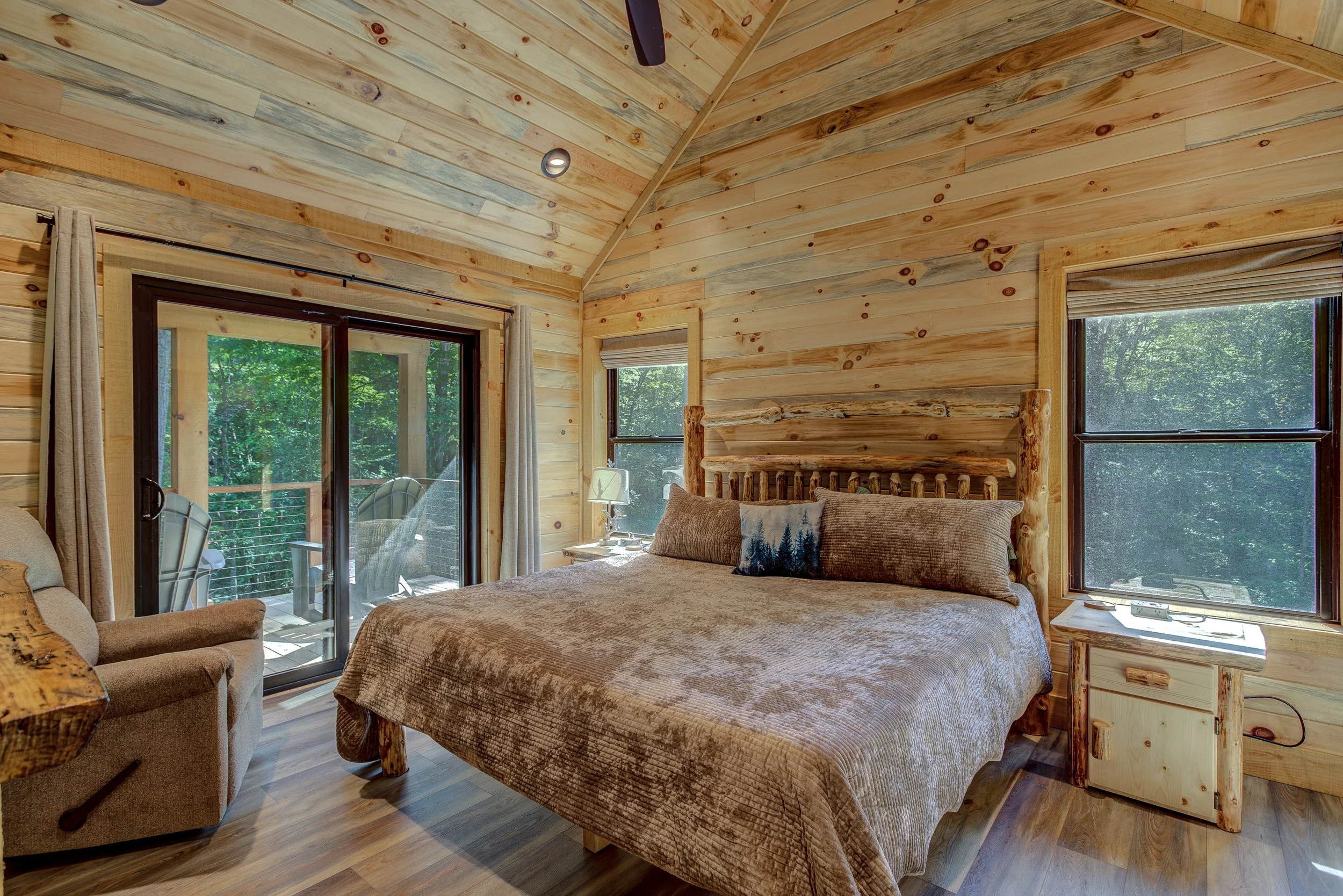 Peaceful cabin bedrooms filled with warmth and character, featuring vaulted natural-wood ceilings, rustic furnishings, large windows, and private sliding doors overlooking the forest. One bedroom also includes a cozy electric fireplace—the perfect re