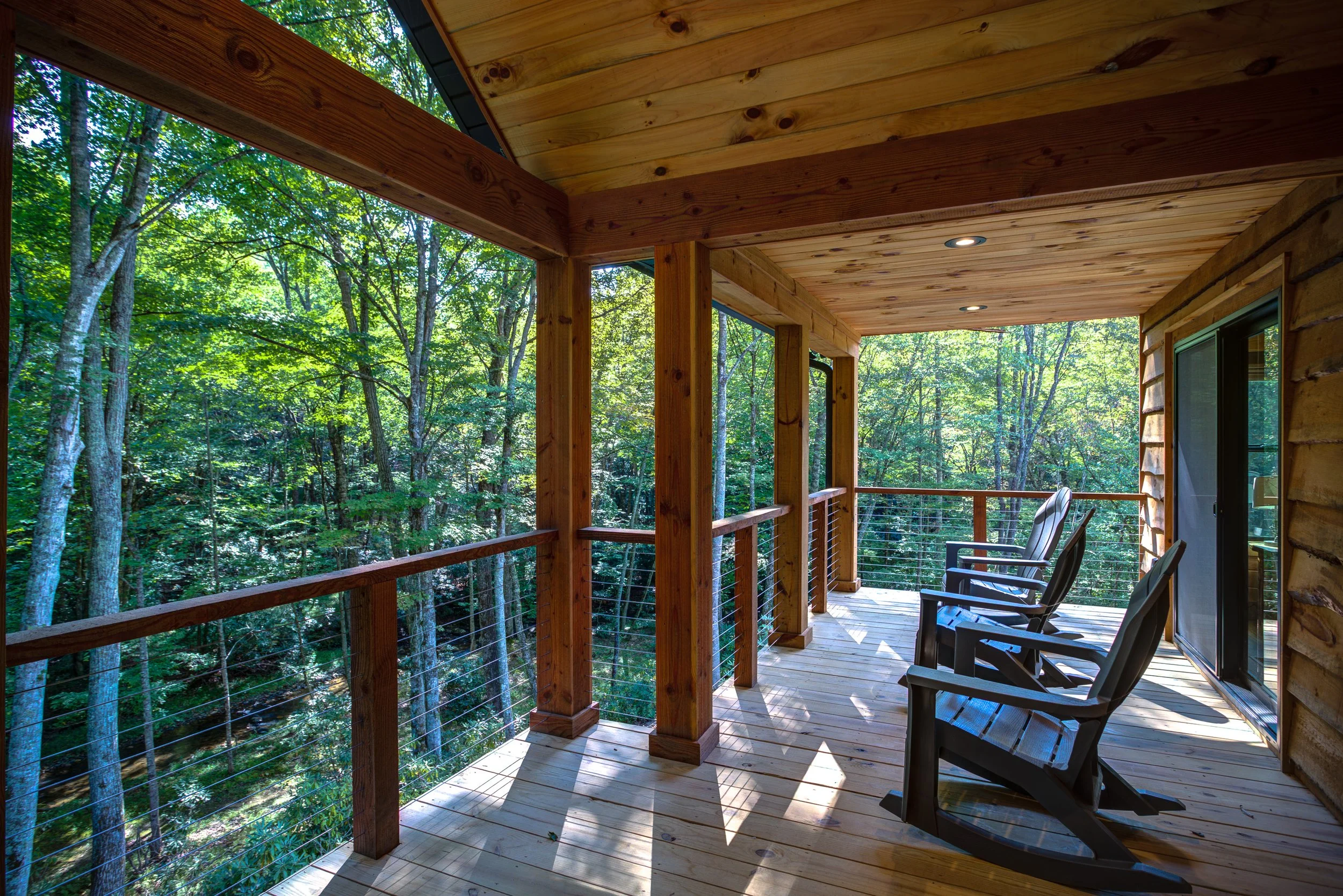 A peaceful covered back porch designed for enjoying the beauty of mountain living. Natural wood ceilings, exposed timber posts, cable railings, recessed lighting, and rustic porch swings create the perfect place to relax while surrounded by the quiet