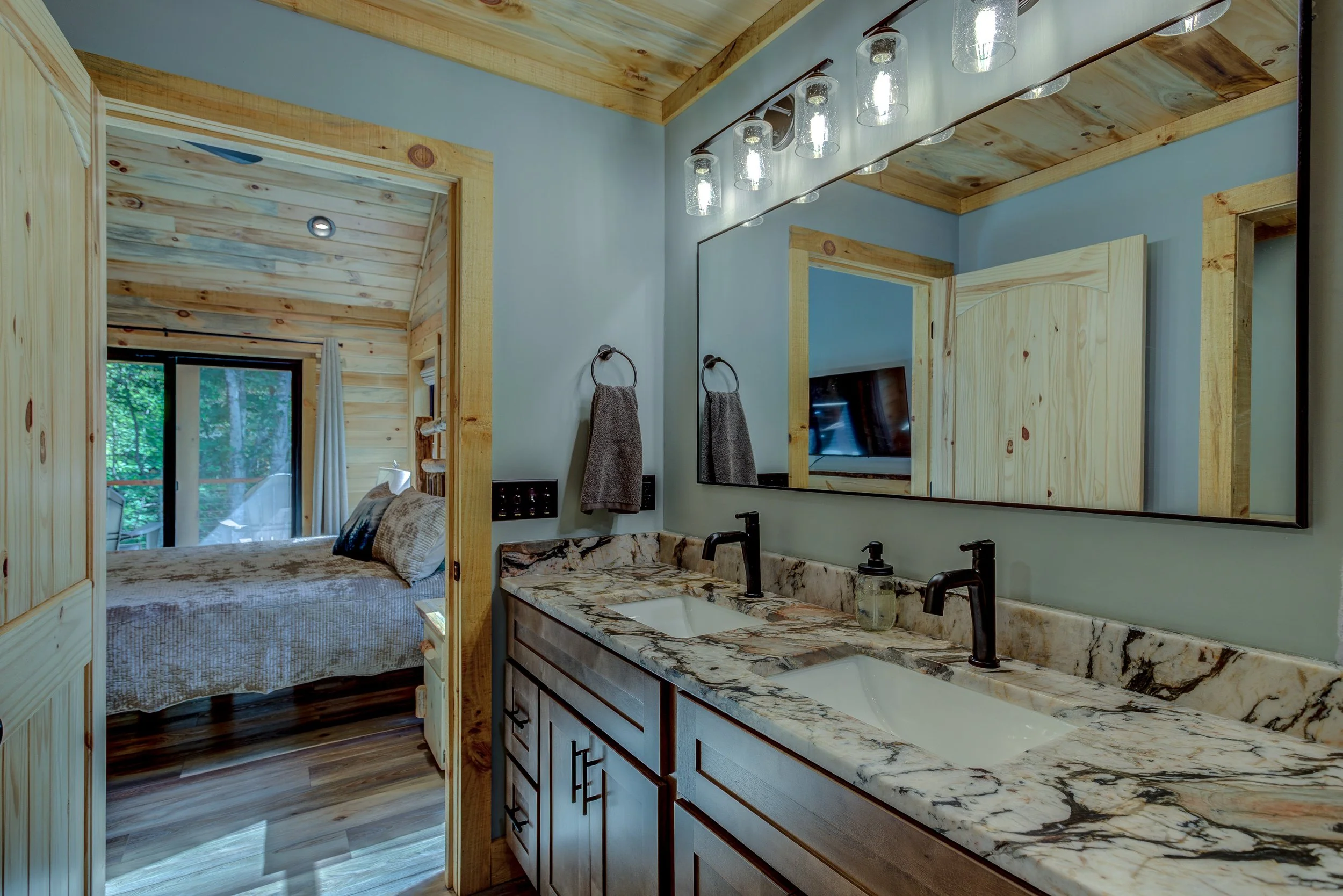 A beautifully finished bathroom featuring a spacious double vanity, natural wood cabinetry, elegant stone-style countertops, and a large walk-in shower surrounded by floor-to-ceiling stone tile. Rustic details and modern finishes create a relaxing, s