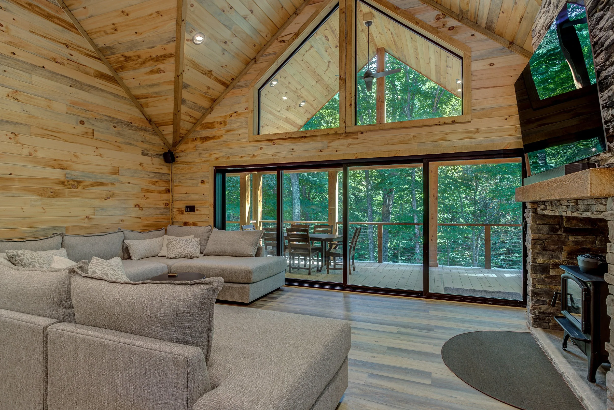 A warm and inviting living room featuring vaulted natural-wood ceilings, an impressive stone fireplace, and comfortable open-concept living. Oversized windows and sliding glass doors fill the space with natural light while showcasing the peaceful for