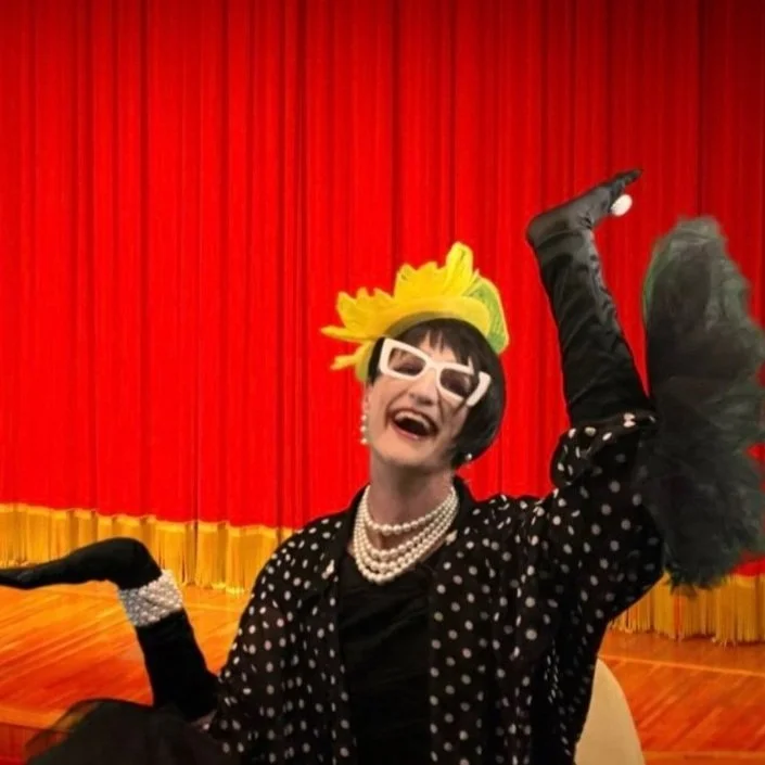 Person in a polka dot outfit with yellow hat and white sunglasses, performing in front of a red curtain.