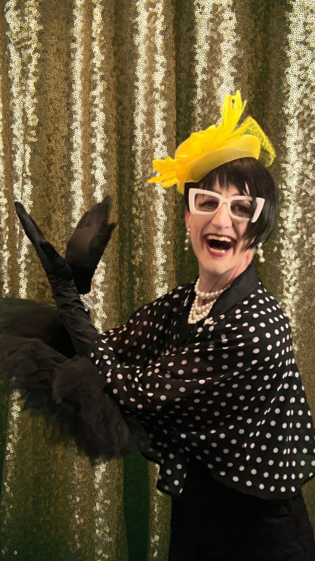 Person wearing polka dot clothing, large white glasses, and yellow feathered hat, posing in front of a golden sequin curtain.