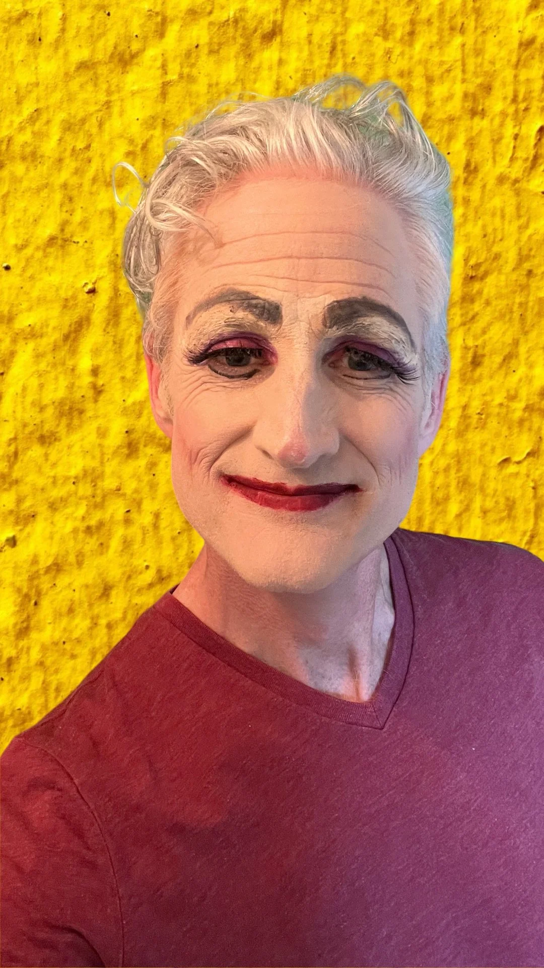 Person with theatrical makeup and white hair against a textured yellow background.