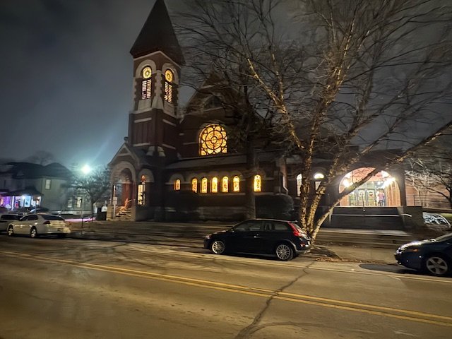Nighttime view of a church with illuminated windows, a large tree in front, and parked cars along the street.
