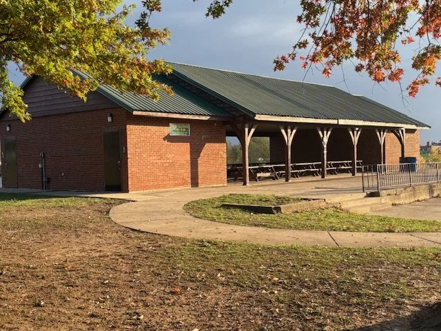 A brick pavilion with a green roof and picnic tables underneath, surrounded by trees with autumn foliage.